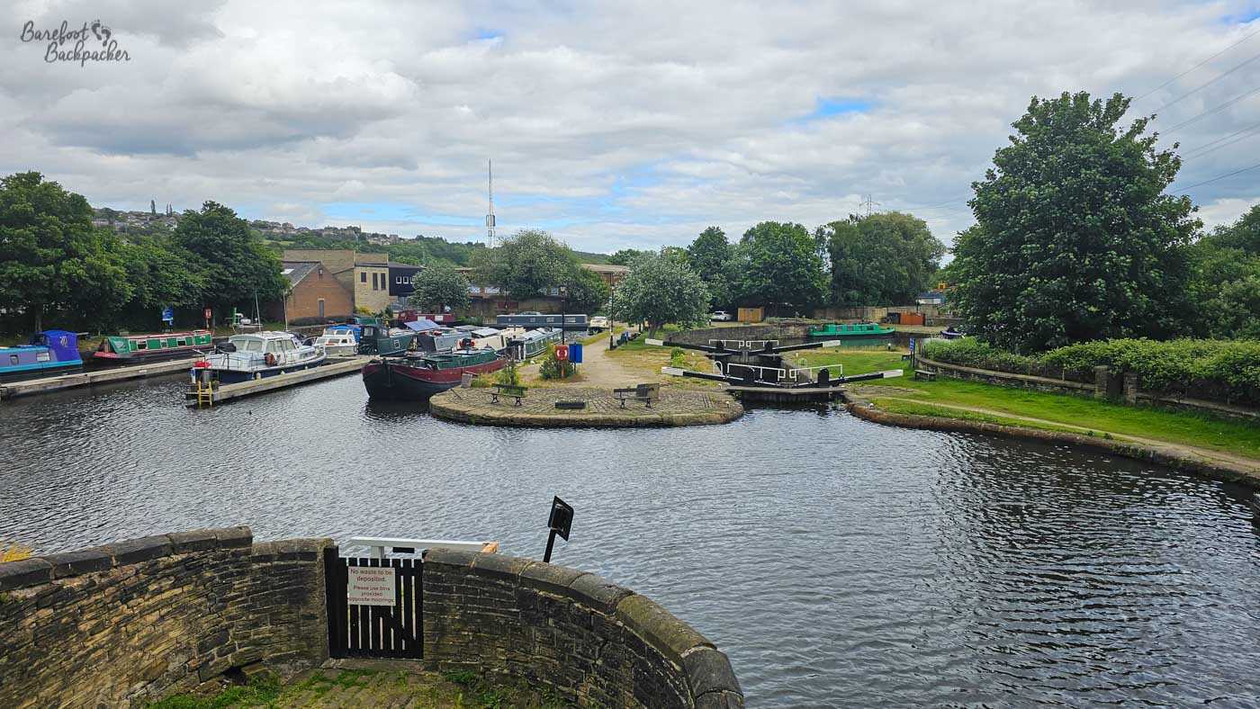 A broad canal basin contains numerous colourful narrowboats and other small boats moored around its edges and central wharf. A small rounded peninsula with benches projects into the water, while black-and-white lock gates and canal structures stand to either side. Trees, low buildings and a hillside neighbourhood form the background beneath a cloudy sky.