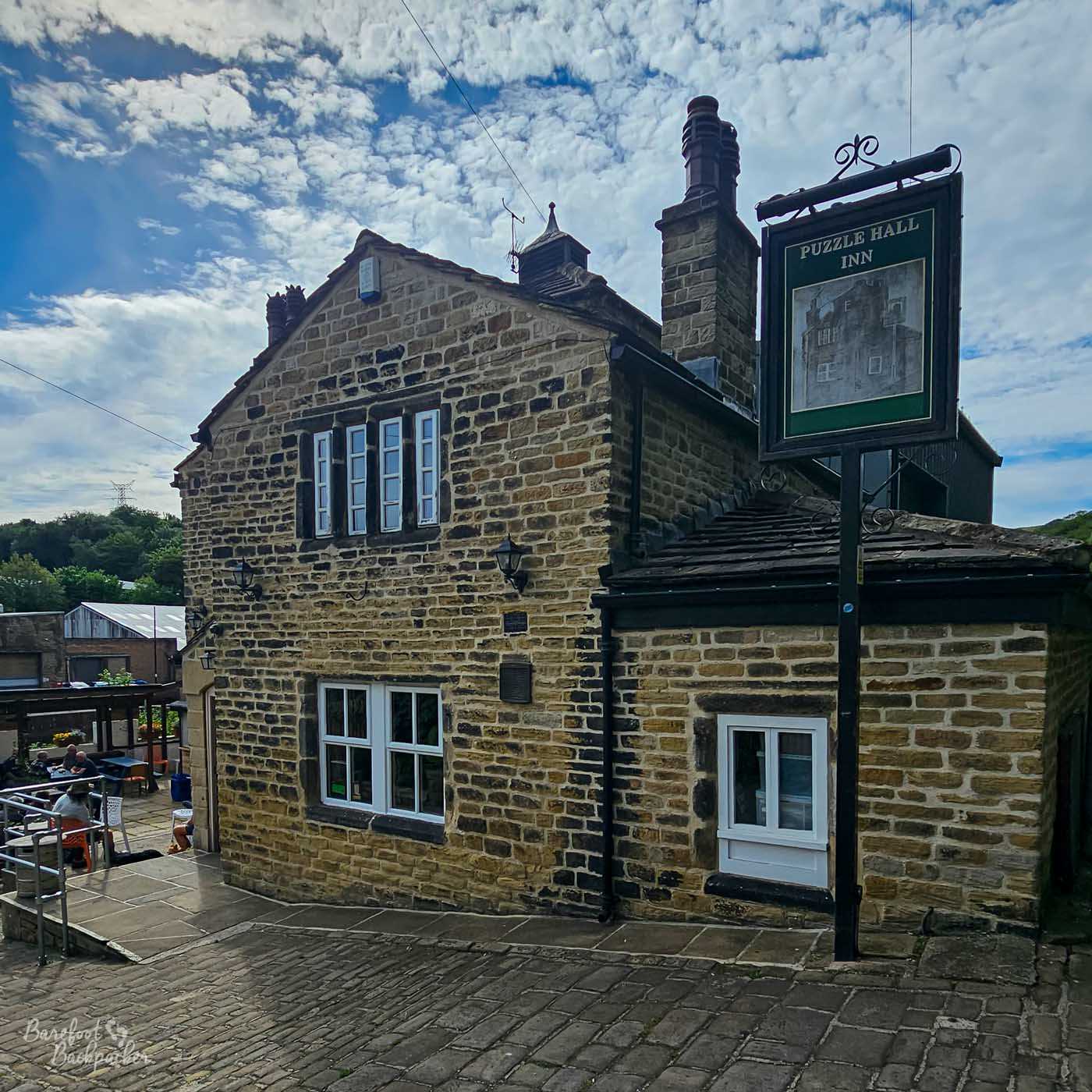 The Puzzle Hall Inn occupies a handsome stone building on a steep cobbled street overlooking the valley below. Its traditional sign hangs beside the pavement, while the pub's outdoor seating stretches onto a sunny terrace at the rear where customers enjoy drinks in the open air. Weathered stone walls, slate roofs and scattered clouds reinforce the historic Yorkshire setting.