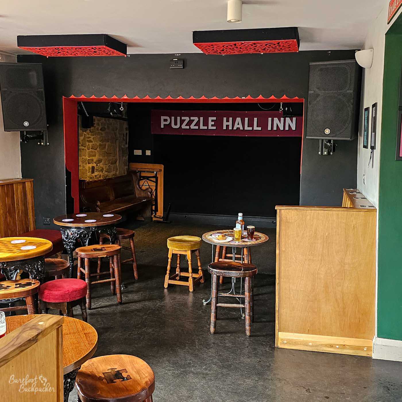 A small performance area sits at one end of the pub, with a simple black stage framed in red beneath a sign reading