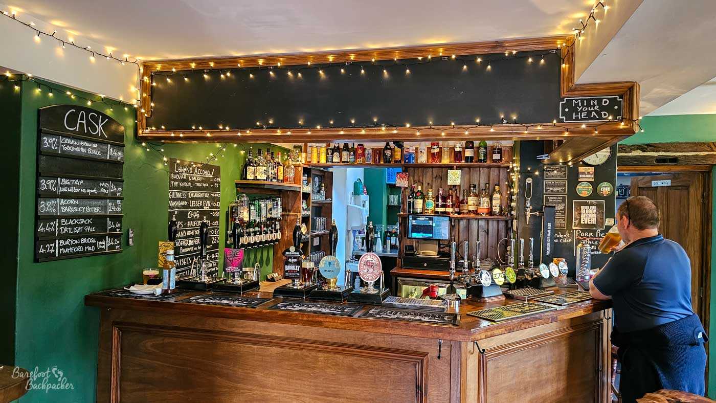 The compact wooden bar is lined with hand pumps offering a wide selection of cask ales, their colourful pump clips standing beneath blackboards listing beers and prices in chalk. Warm fairy lights frame the bar area, while shelves of spirits fill the back wall and a customer leans casually against the counter enjoying a pint. Dark green walls, polished timber and traditional pub décor create the cosy atmosphere of a well-established real ale pub.