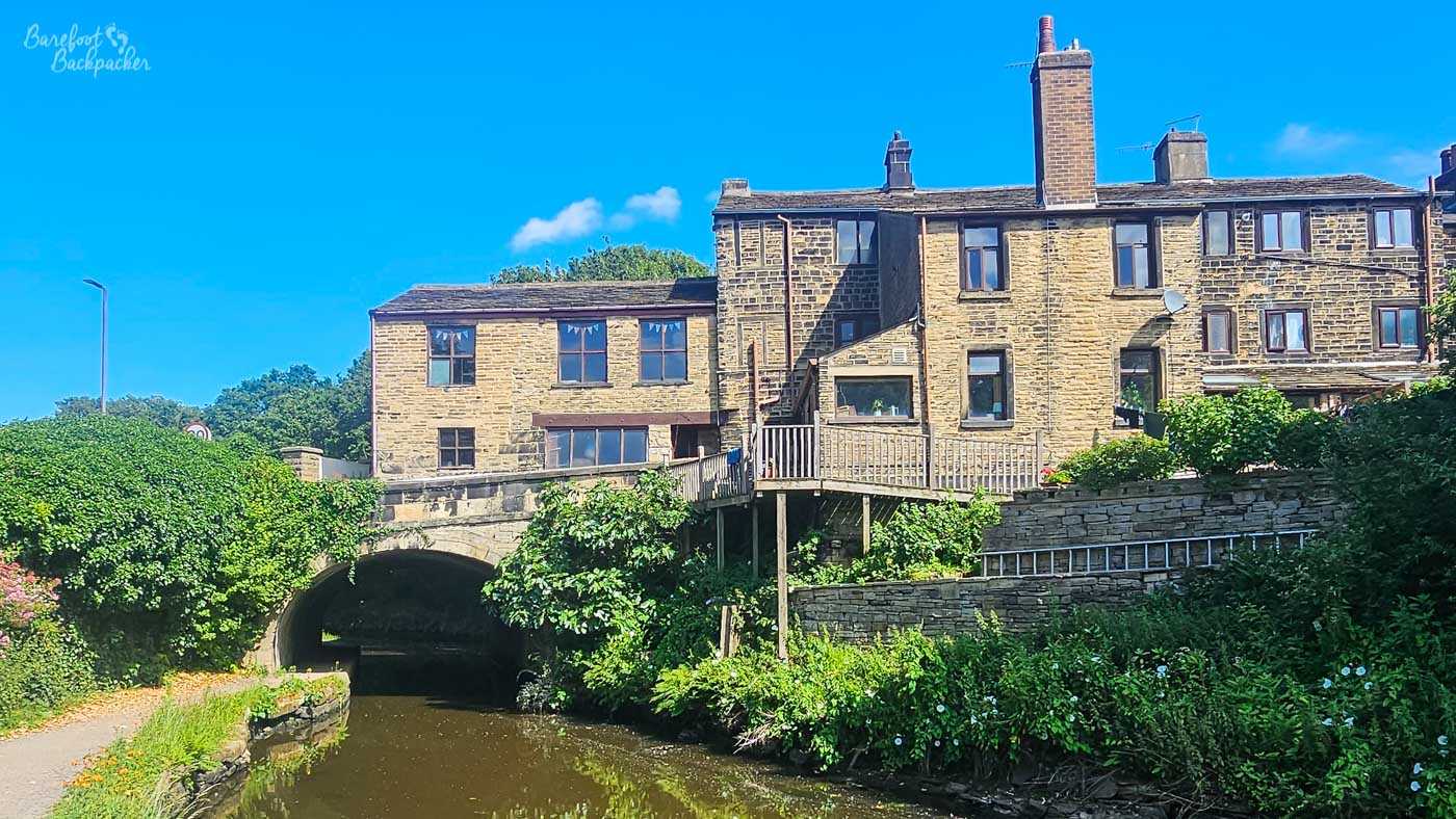 The canal curves beneath a low stone bridge with traditional Yorkshire stone houses built closely above and beside it. Wooden balconies and terraces project from the rear of the buildings, while thick greenery covers much of the canal bank and spills around the bridge. Bright sunshine illuminates the golden stonework beneath a vivid blue sky.