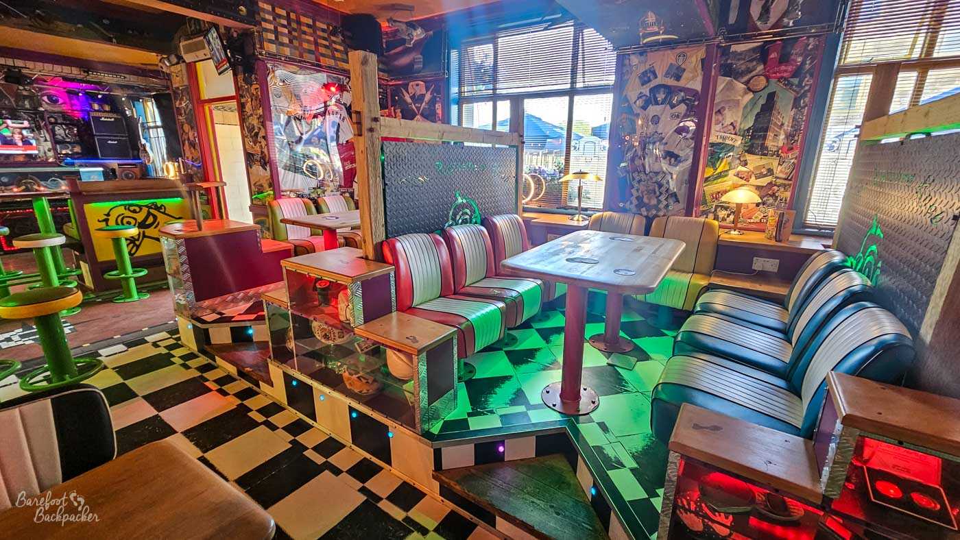 Inside The Polished Knob, colourful retro booth seating surrounds raised tables on a black-and-white chequered floor. Neon lighting, vintage posters, music memorabilia and quirky decorations fill every wall, creating a vibrant, eclectic space with a distinctly alternative atmosphere. Large windows allow natural light to mix with the coloured interior lighting.