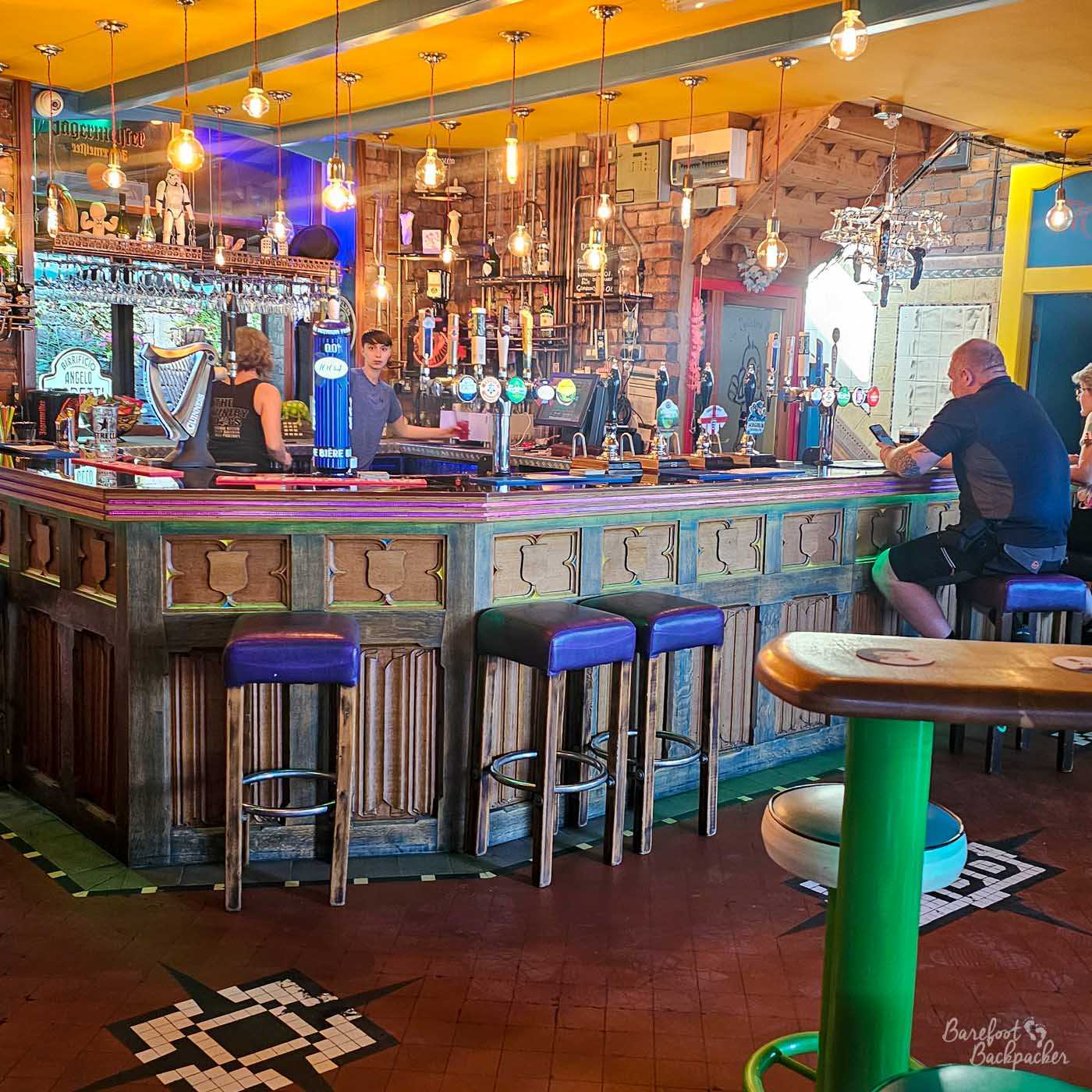 The large central bar at The Polished Knob is surrounded by tall stools beneath rows of warm hanging filament bulbs. Numerous hand pumps line the counter while shelves of bottles and glassware fill the back bar. A handful of customers sit chatting or enjoying drinks, giving the spacious room a relaxed but lively feel.