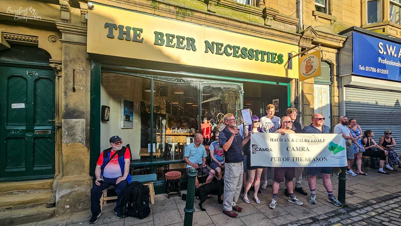 A group of CAMRA members gather outside Beer Necessities during an award presentation, holding a large banner recognising the pub as Halifax & Calderdale CAMRA Pub of the Season. Several people watch from the pavement while others sit outside with drinks in the warm evening sunshine. The pub's wide picture window reflects the busy street behind the ceremony.