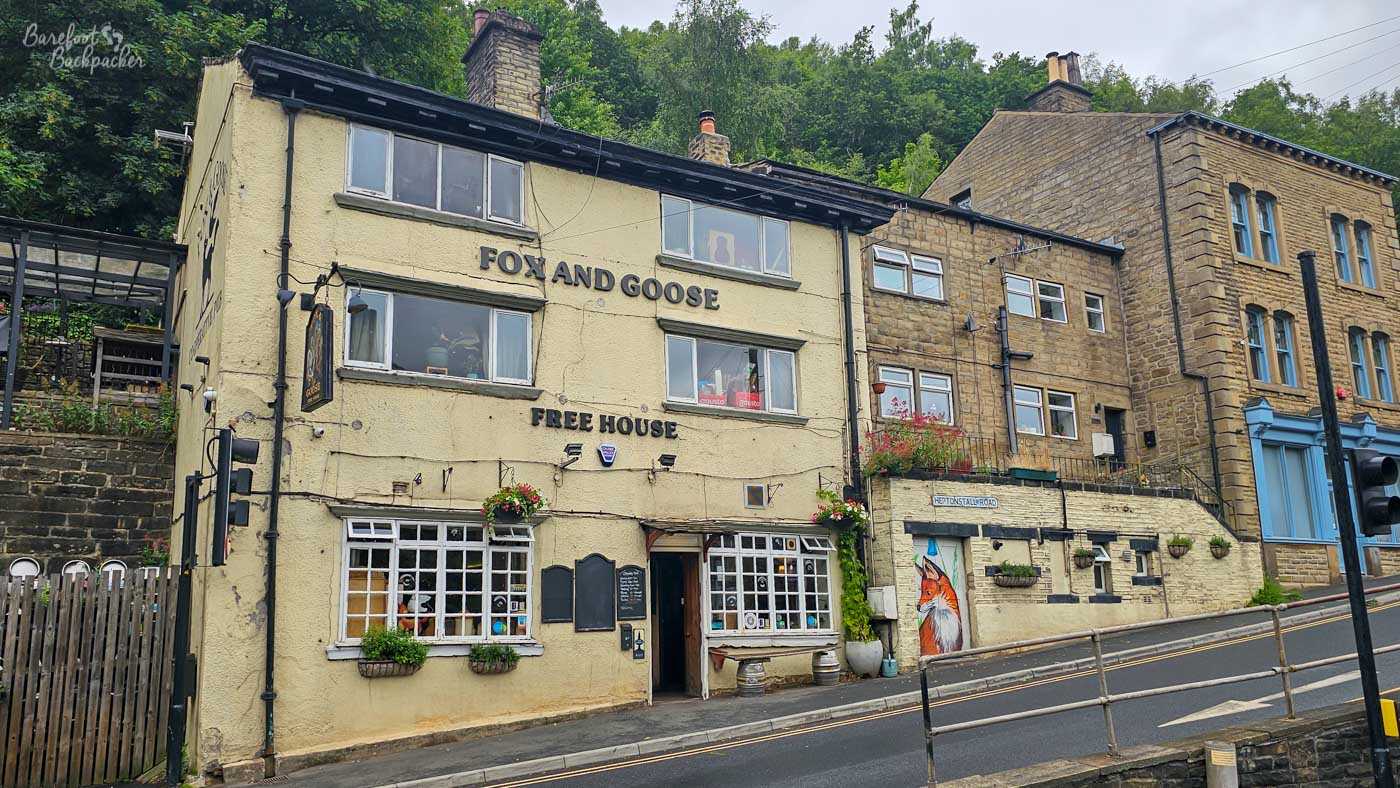 Exterior of the Fox and Goose, a traditional free house on a sloping street in Hebden Bridge. The pale cream, slightly weathered three-storey pub has large black lettering reading FOX AND GOOSE and FREE HOUSE across its frontage, with white-framed windows, hanging flower baskets and an open central doorway. It sits among adjoining stone buildings at the foot of a steep, densely wooded hillside, while the road and metal pedestrian railings descend across the foreground.