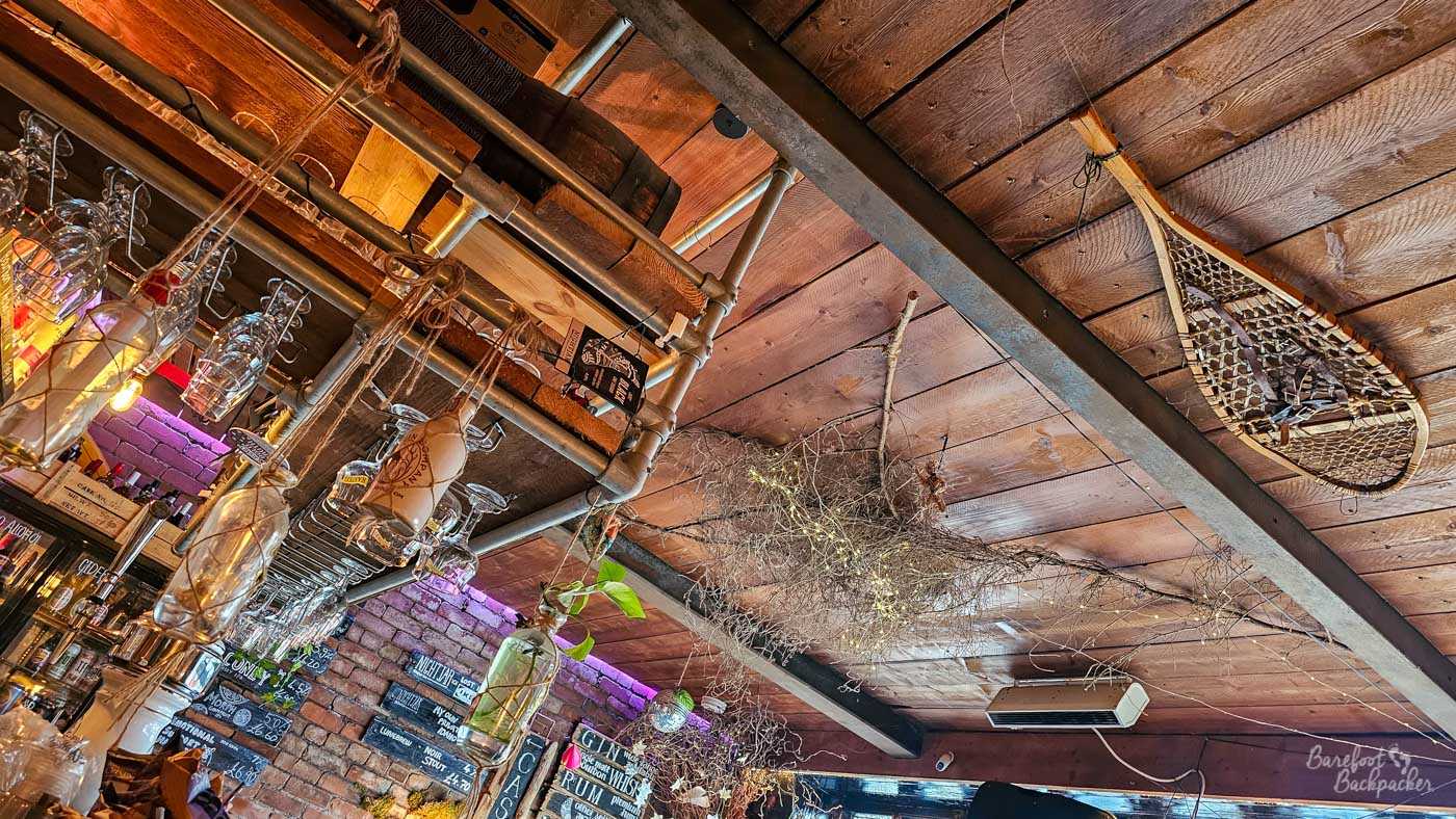 Looking upwards at the unusually decorated wooden ceiling inside Nightjar in Hebden Bridge. Heavy dark beams and exposed metal pipes cross the timber boards, with bottles and glasses suspended above the bar on the left. Tangled bare branches threaded with tiny lights hang across the centre of the ceiling, while an old-fashioned wooden-framed snowshoe is mounted overhead on the right. The jumble of industrial fittings, reclaimed objects and rustic decorations continues the bar's quirky interior style.