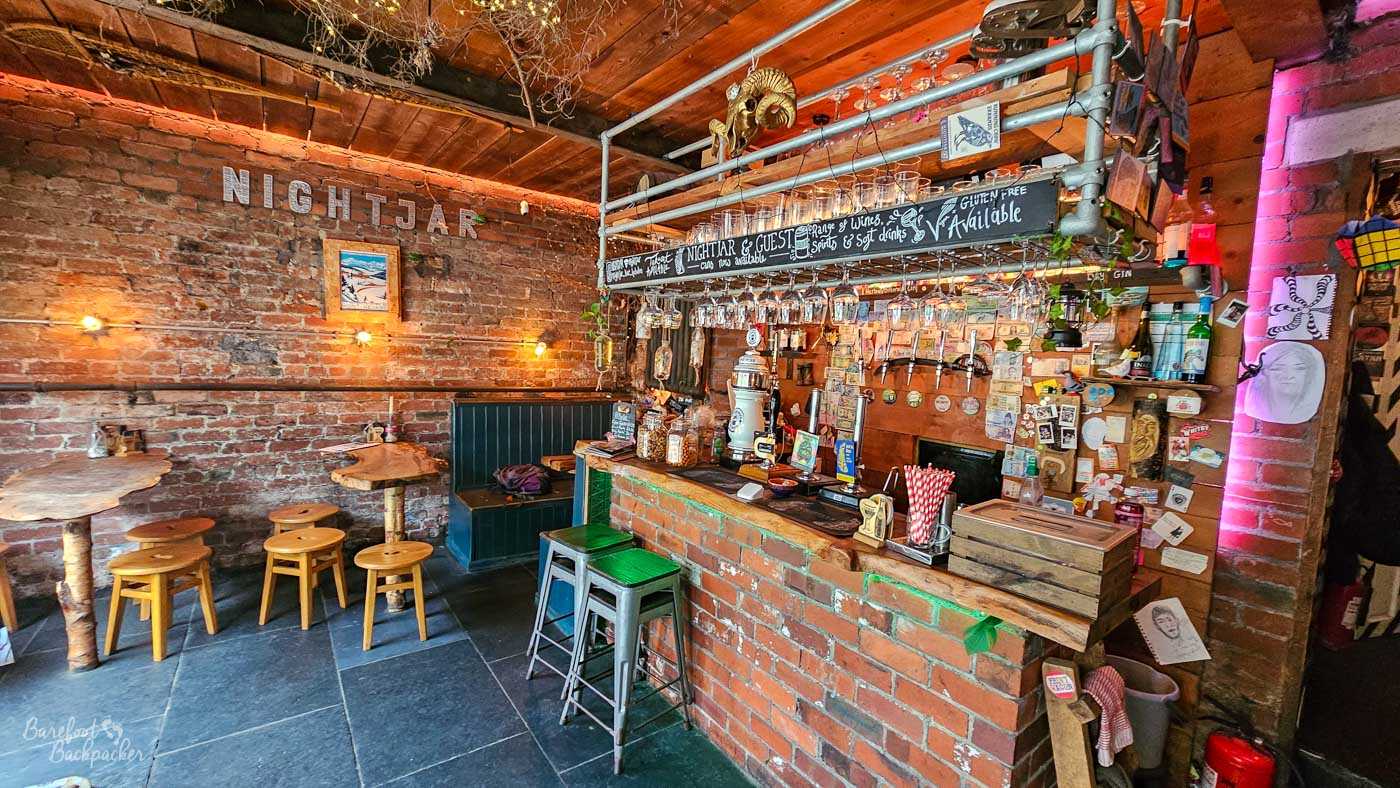 Rustic interior of Nightjar in Hebden Bridge, centred on a compact bar built from exposed red brick and rough-edged timber. Beer taps line the brick wall behind it beneath racks of hanging glasses and handwritten signs advertising drinks, while postcards, stickers and assorted small objects cover much of the surrounding brickwork. Wooden stools and irregular natural-edged tables sit against the exposed-brick walls, with warm lighting and visible pipes adding to the deliberately rough, eclectic appearance.