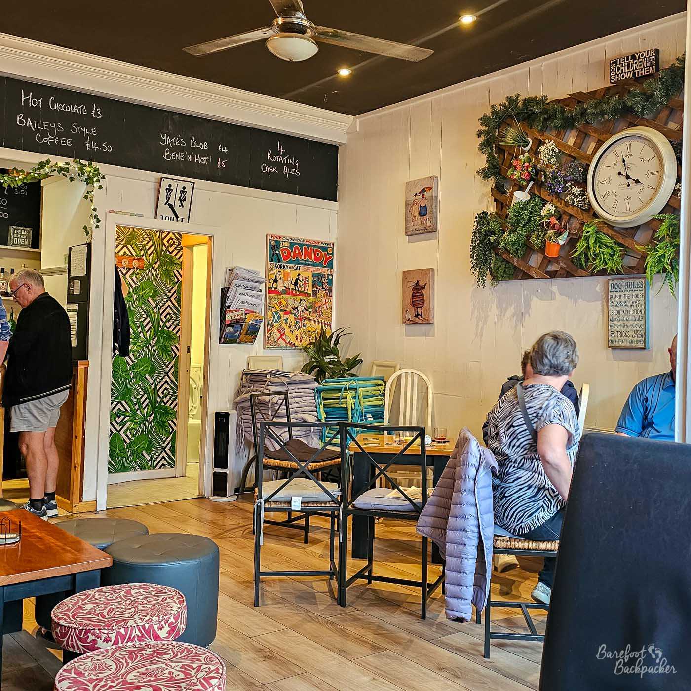 The Alehouse has a bright, welcoming interior with pale wooden floors, black-framed tables and a mixture of chairs and upholstered seating. A large decorative clock surrounded by greenery hangs on one wall, while handwritten chalkboards above advertise cask ales, coffee and hot drinks. Several customers sit chatting over drinks in the cosy café-style setting.