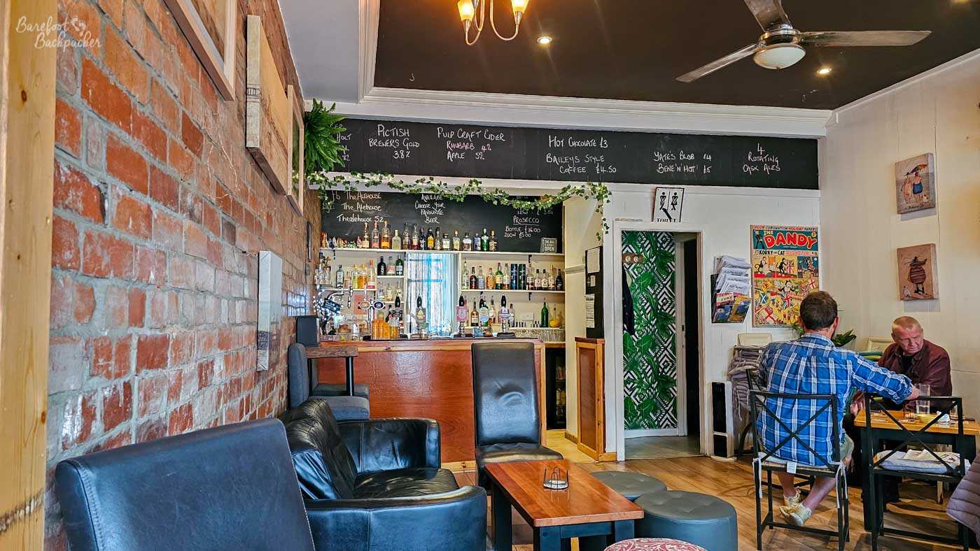 Viewed from the seating area, the compact wooden bar at The Alehouse stands beneath long chalkboards listing beers, ciders and hot drinks. Shelves behind the counter display bottles and spirits, while exposed brickwork, leather armchairs and wooden tables create a relaxed, comfortable atmosphere. A couple of customers enjoy quiet conversation nearby.