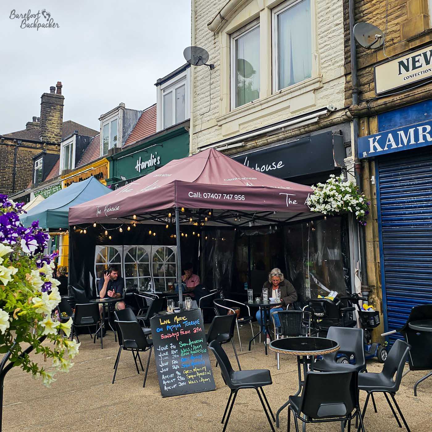 Outdoor tables sit beneath a large maroon canopy outside The Alehouse on Todmorden's main street. Several customers enjoy drinks despite the grey weather, while a chalkboard advertises upcoming live music performances. Hanging flower baskets brighten the frontage, giving the pavement seating an inviting feel.
