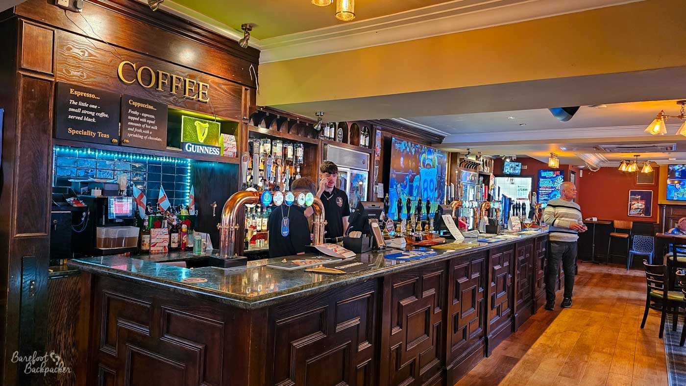 A long traditional wooden bar stretches through The White Hart beneath warm lighting and dark timber panelling. Staff stand behind rows of hand pumps while customers chat nearby, with television screens showing sport further along the room. Decorative Guinness signs, polished brass beer fonts and dark wood furnishings give the pub a classic, contemporary feel.