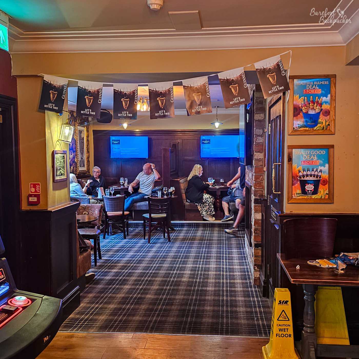 Beyond an archway, customers sit in comfortable booths beneath strings of Guinness bunting in a carpeted lounge area. Dark wood panelling, framed pictures and soft lighting create a relaxed atmosphere, while television screens mounted on the far wall suggest the pub regularly shows live sport. A yellow