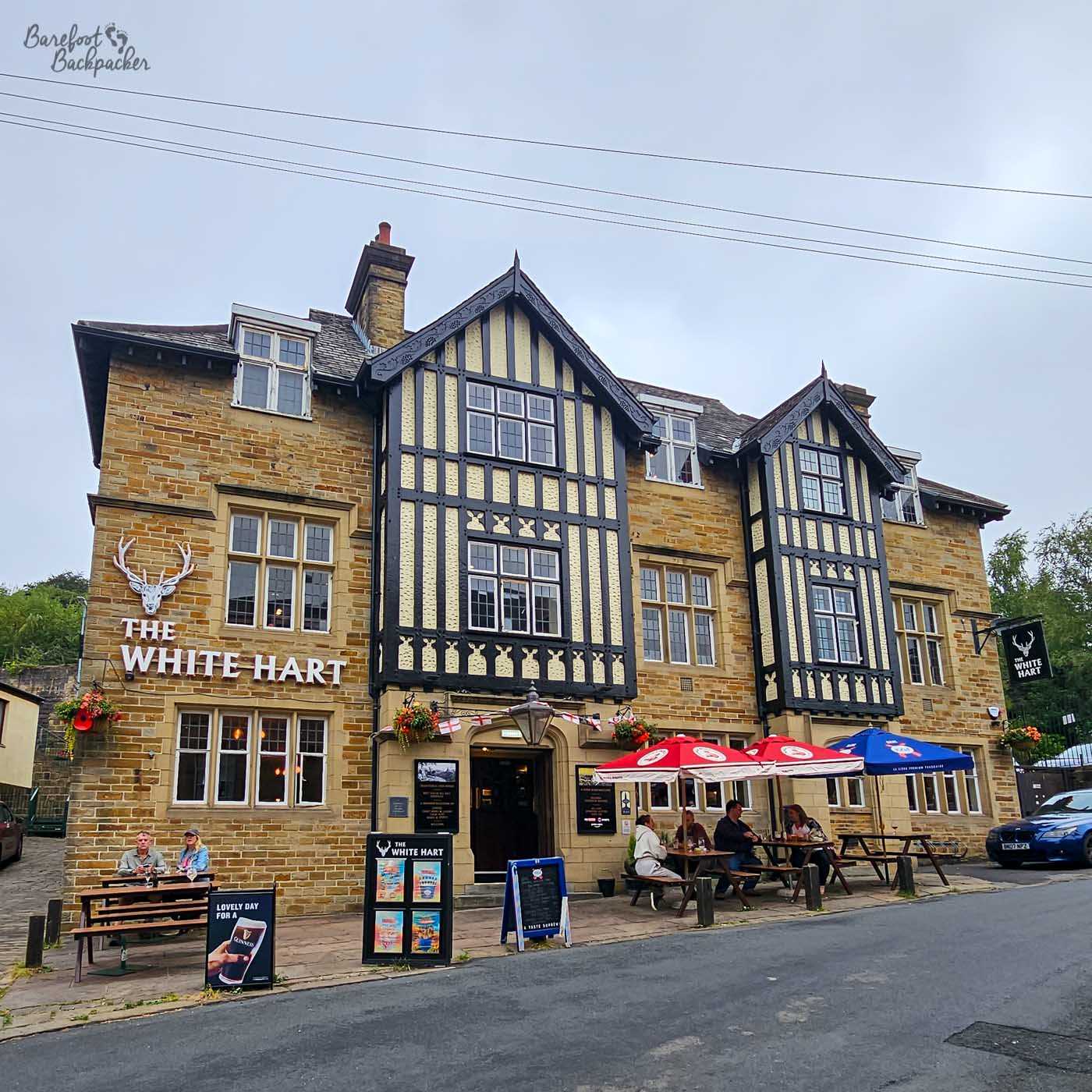 The White Hart occupies an impressive stone building with striking black-and-white timbered gables above its entrance. Outdoor picnic benches with red and blue umbrellas line the pavement, where several customers enjoy drinks outside. Hanging baskets and traditional pub signage complete the historic frontage on an otherwise quiet street.