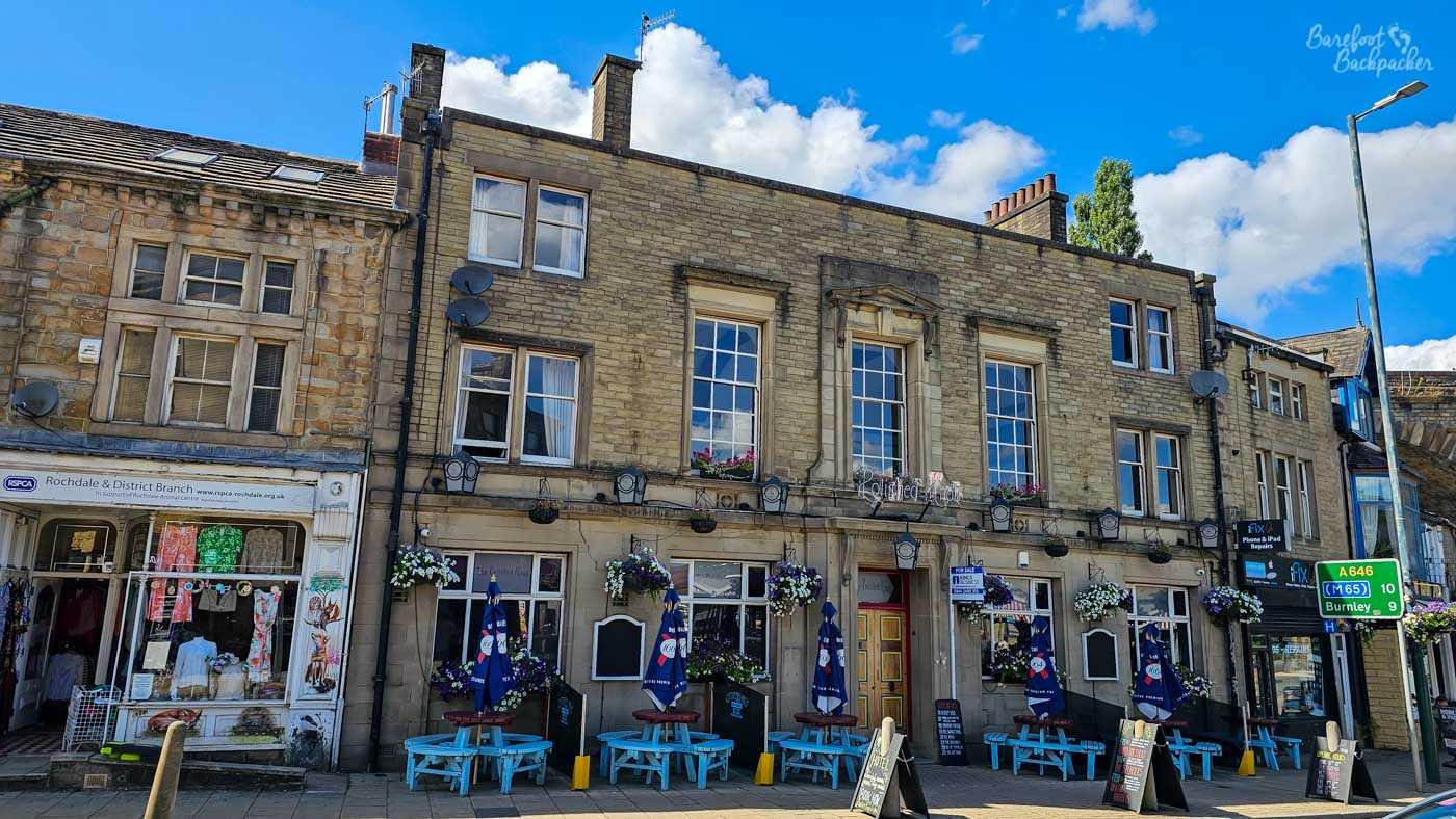 The broad stone frontage of The Polished Knob occupies a prominent position on Todmorden's high street. Hanging flower baskets decorate the building, while bright blue picnic benches and folded parasols line the pavement outside. Surrounded by neighbouring shops, the pub combines traditional Yorkshire stone architecture with a lively town-centre presence.
