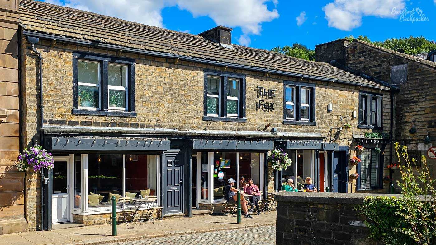 The stone-built frontage of The Fox pub in Todmorden sits beside a cobbled street on a sunny afternoon. Hanging flower baskets brighten the pale stone building, while large front windows reveal glimpses of the cosy interior. Several people relax at small pavement tables outside, enjoying drinks in the sunshine beneath a bright blue sky dotted with white clouds.