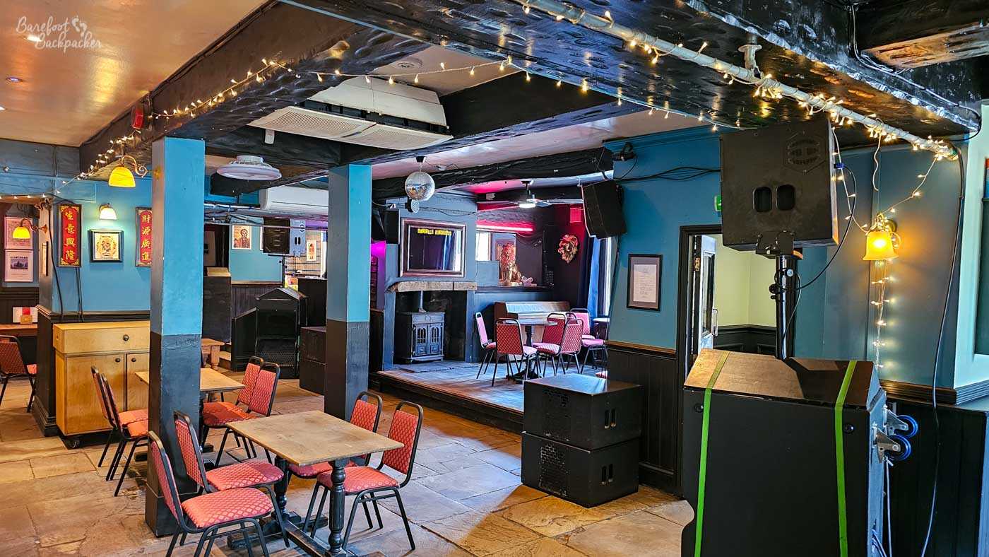 The main room of The Golden Lion combines a traditional pub with a live music venue, featuring small tables with red upholstered chairs facing a low stage. Large speakers, fairy lights and a disco ball hint at regular gigs, while blue-painted pillars divide the open space. A black wood-burning stove sits at the back of the stage beneath soft coloured lighting.