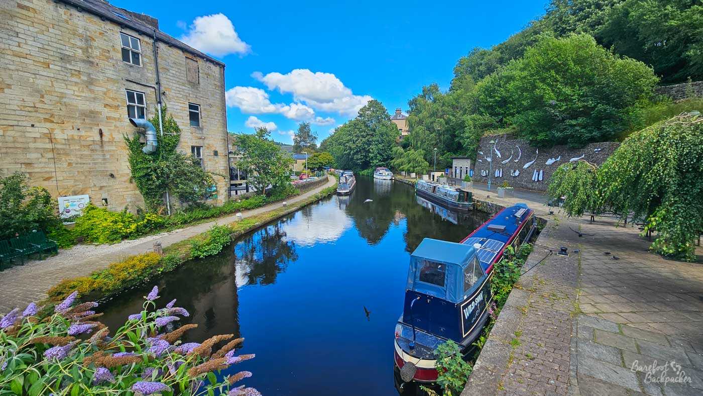 Several narrowboats are moored along a wide section of canal, including a dark blue boat in the foreground beside a paved waterside area. An old pale-stone building stands on the opposite bank, while dense trees rise behind the canal on the right. The still water reflects the boats, trees and vivid blue summer sky, and a large mural decorates the retaining wall beside the moorings.