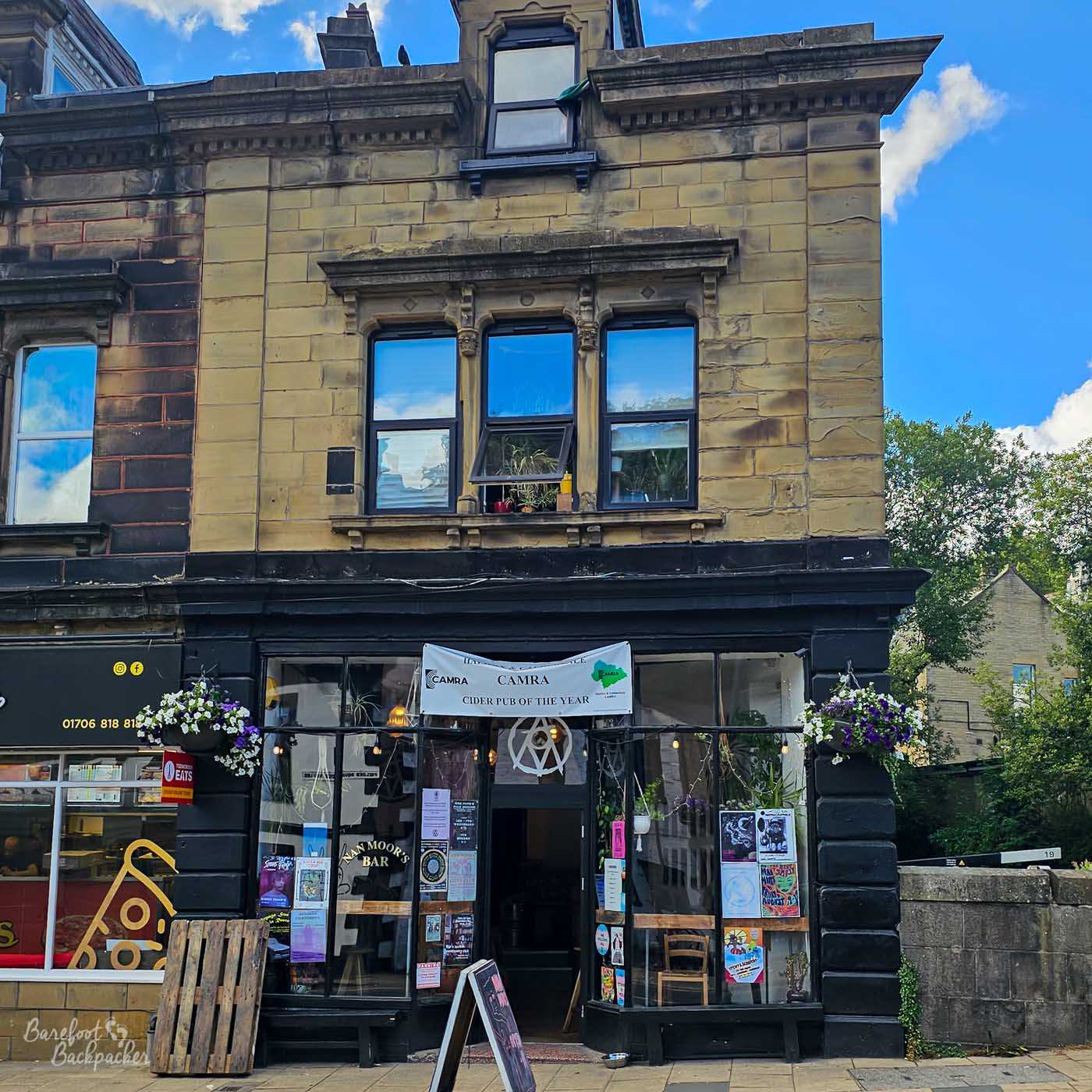 The narrow Victorian frontage of Nan Moor's Bar rises above a street-level entrance, its black-painted ground floor contrasting with the pale sandstone upper storeys. Posters and notices fill the windows, while a CAMRA Cider Pub of the Year banner hangs above the doorway. Hanging baskets add colour to the entrance, giving the independent pub an eclectic and welcoming appearance.
