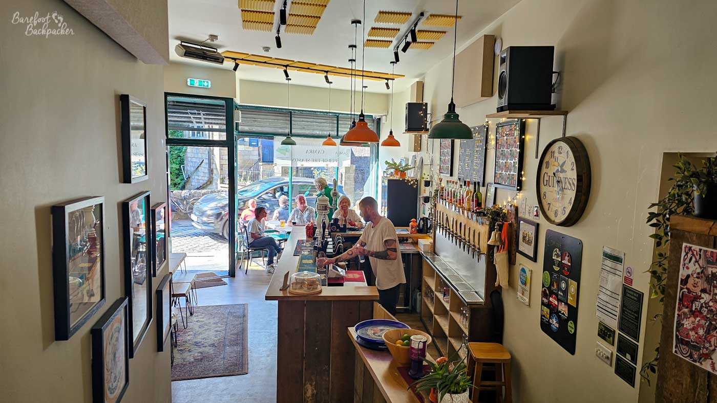 Viewed from a raised step, Beer Necessities has a long, narrow layout centred around a simple wooden bar. A bartender serves drinks beneath colourful pendant lights while rows of beer taps line one wall alongside a large Guinness clock. Through the open frontage, customers can be seen enjoying drinks at tables outside on the pavement.