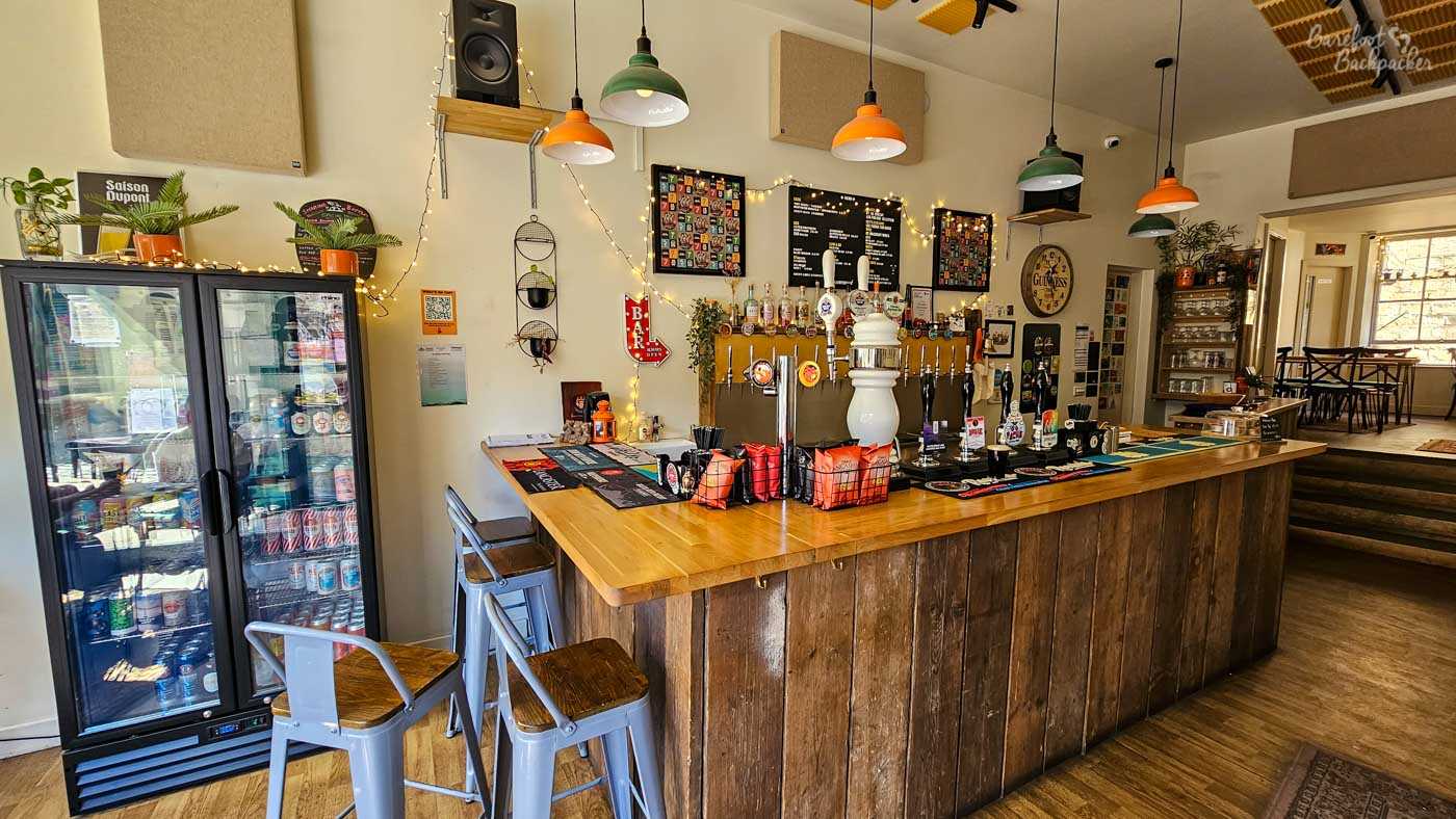 The compact wooden bar at Beer Necessities is lined with hand pumps serving a wide selection of beers, while chilled cans and bottles fill a glass-fronted fridge beside it. Fairy lights, colourful pump clips and framed beer memorabilia decorate the walls, giving the modern micropub a bright yet informal atmosphere. Pendant lights hang from the high ceiling above the polished wooden counter.