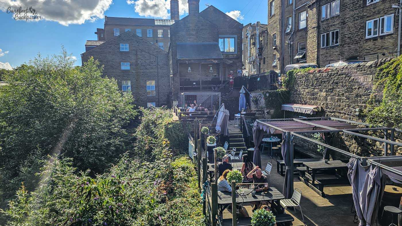 The terraced beer garden behind the Turks Head cascades down the hillside through several levels of wooden decking overlooking a densely wooded valley. Customers sit at outdoor tables beneath folded parasols enjoying the afternoon sunshine, while stone retaining walls and neighbouring buildings frame the secluded space. Despite being close to the town centre, the garden feels unexpectedly peaceful and sheltered among the greenery.