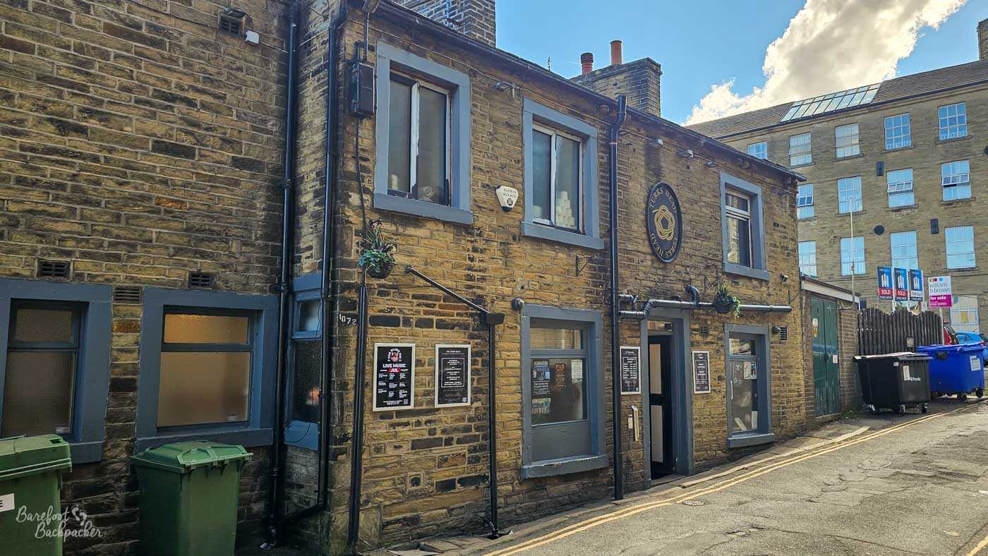 The Turks Head occupies a modest stone building tucked away along a narrow back street between larger former mill buildings and modern apartments. Hanging baskets soften the traditional Yorkshire stone façade, while posters beside the doorway advertise forthcoming live music events. Its understated appearance makes it feel like one of those pubs discovered by wandering away from the main road.