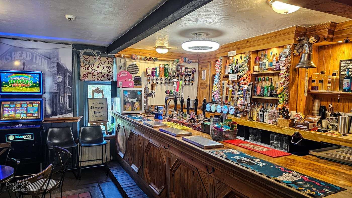 A traditional wooden bar stretches along one side of the compact interior, its polished counter backed by shelves of spirits, rows of hand pumps and colourful beer mats. Warm timber panelling, low ceilings and soft lighting create a cosy atmosphere, while a fruit machine and small seating area occupy one corner of the room. The space feels welcoming and unmistakably like a classic local pub that has evolved over many years rather than being designed all at once.
