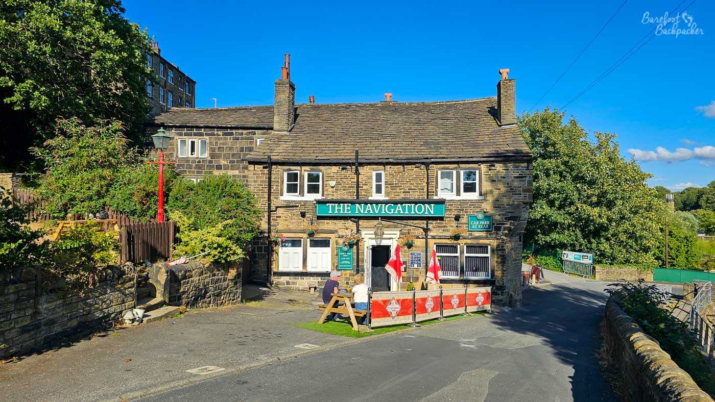 The Navigation occupies a traditional two-storey Yorkshire stone building standing beside the road close to the canal. A large green sign stretches across the frontage above the entrance, while a small outdoor seating area with picnic benches and red brewery umbrellas sits at the front. Mature trees, stone walls and neighbouring cottages frame the pub, giving it the feel of a long-established local.