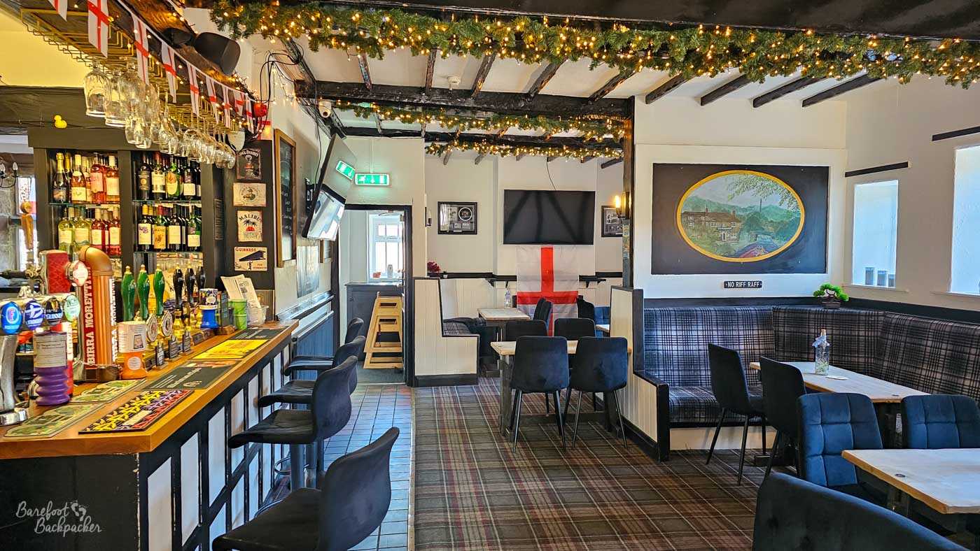 The main bar blends traditional pub features with modern furnishings, including exposed ceiling beams decorated with garlands and fairy lights above a patterned carpet. A compact wooden bar stocked with cask ales and spirits runs along one wall, while booth seating, televisions and framed artwork create a comfortable, welcoming atmosphere. Black-and-white décor contrasts with warm timber details throughout the room.