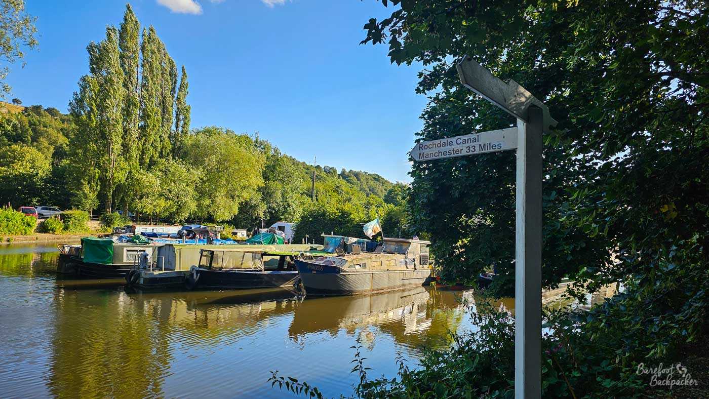 A white wooden signpost beside the canal reads Rochdale Canal, Manchester 33 Miles, pointing to the left. Several narrowboats are clustered on the water behind it, surrounded by dense green trees and vegetation. Bright sunshine reflects the boats and greenery in the gently rippled canal.