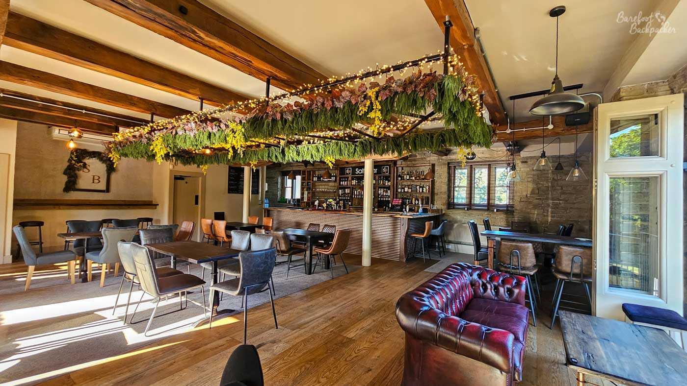 A large open lounge combines exposed stone walls and heavy timber beams with contemporary furniture and lighting. Suspended greenery and decorative foliage hang above the seating area, while sunlight streams through the windows towards a modern wooden bar at the far end of the room. Leather chairs, dining tables and open floor space give the venue a bright, airy feel.