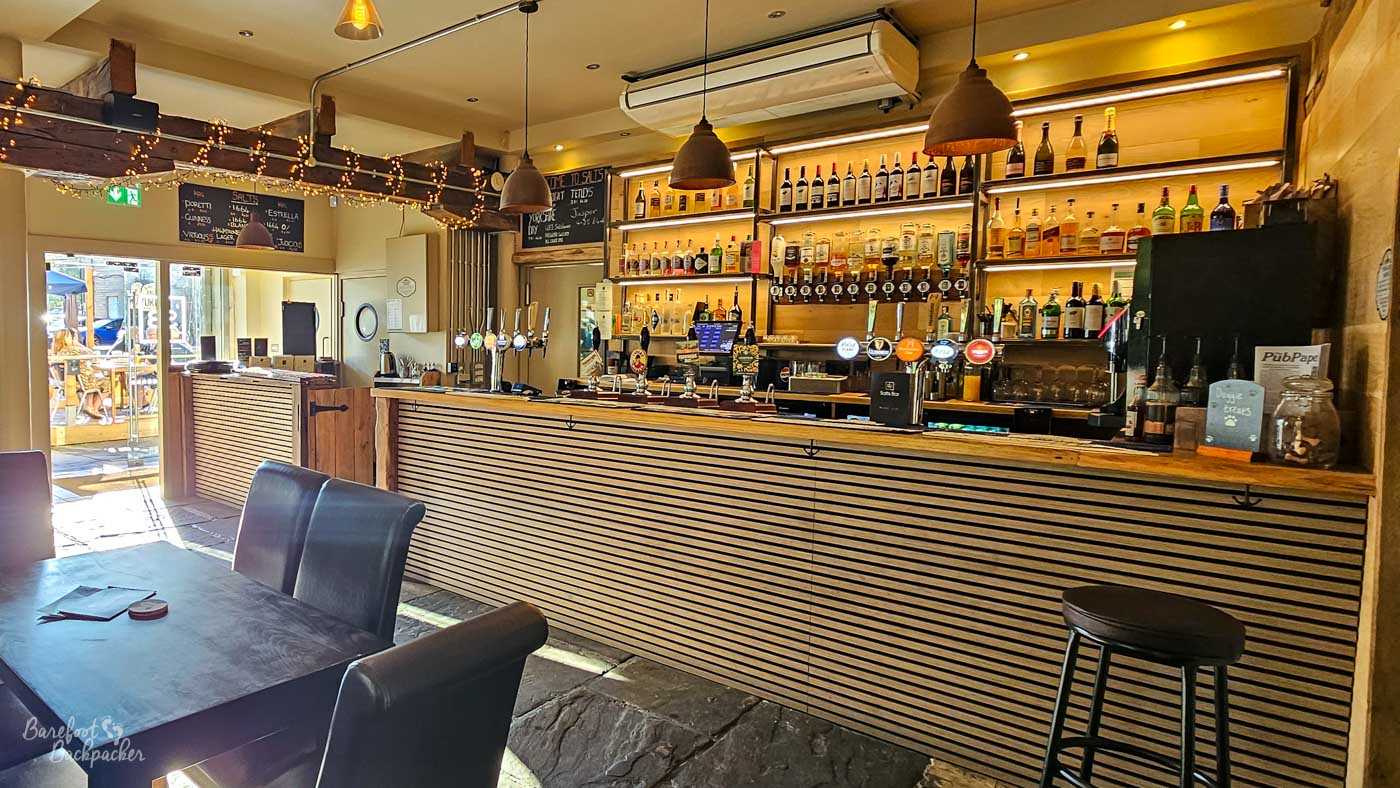 The modern timber-fronted bar is lined with hand pumps, spirits and wine displayed on illuminated shelving behind the counter. Pendant lights hang from the ceiling beams while sunlight pours through the open doorway leading out to the beer garden. Stone flooring and exposed timber create a contemporary interpretation of a traditional Yorkshire pub interior.
