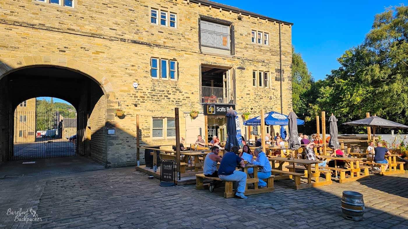 Salts Bar occupies part of an impressive converted stone mill building beside a large arched entrance leading into the courtyard beyond. Wooden picnic benches fill the spacious outdoor seating area where customers enjoy food and drinks beneath branded parasols in the sunshine. The historic industrial architecture provides an impressive backdrop while retaining its original stone character.