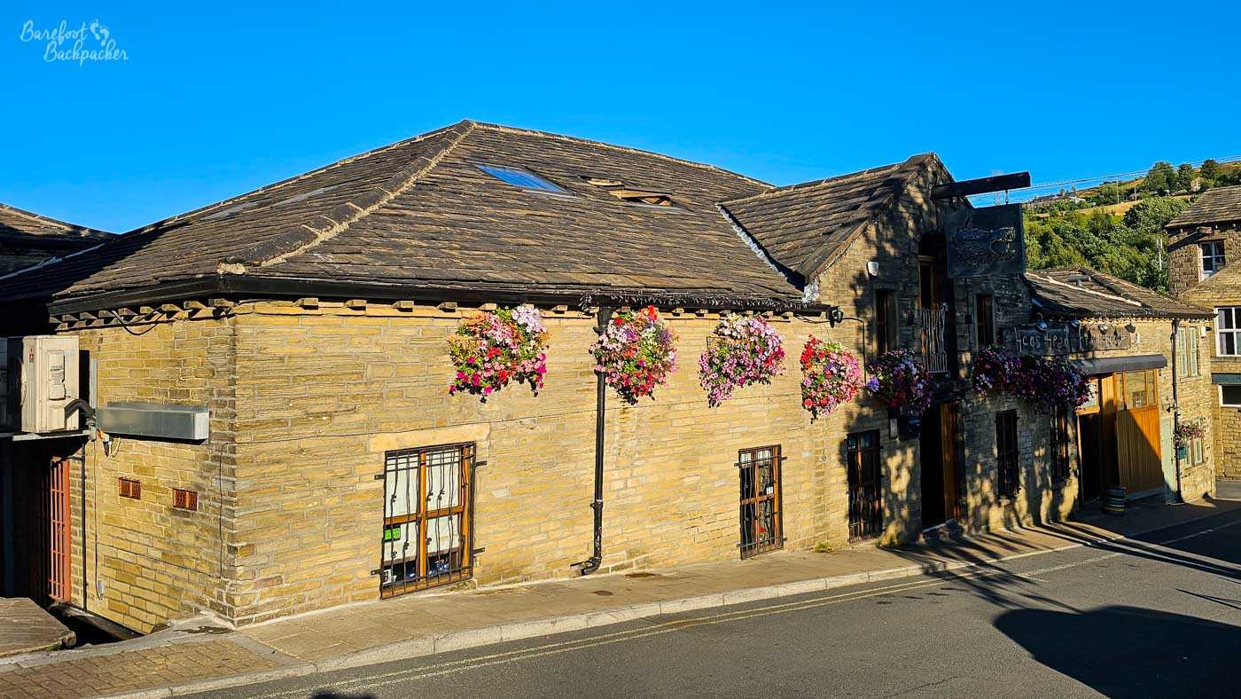 The Hogs Head Brewhouse occupies a long, low stone building beside the road, its traditional Yorkshire stone walls warmed by late afternoon sunshine. Colourful hanging baskets overflowing with pink and purple flowers brighten the frontage, while the dark slate roof and weathered stonework reflect the town's industrial heritage. The pub sits against a backdrop of steep green hills rising beyond the rooftops.
