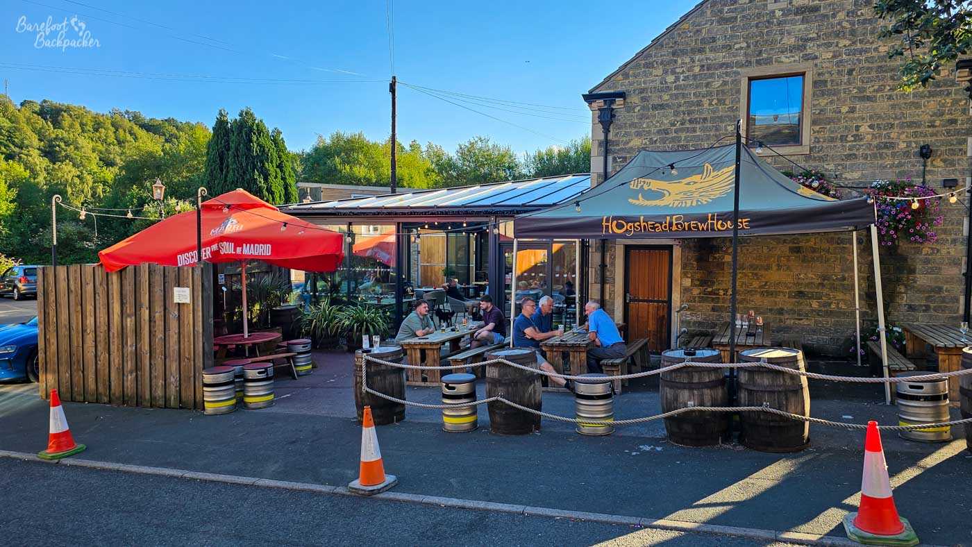 The outdoor seating area wraps around one side of the brewery, with wooden picnic tables beneath branded canopies and large red umbrellas. Groups of customers chat over drinks while decorative barrel planters, rope barriers and strings of lights create an informal courtyard atmosphere. Behind them, the brewery's modern glass extension contrasts with the older stone buildings surrounding it.