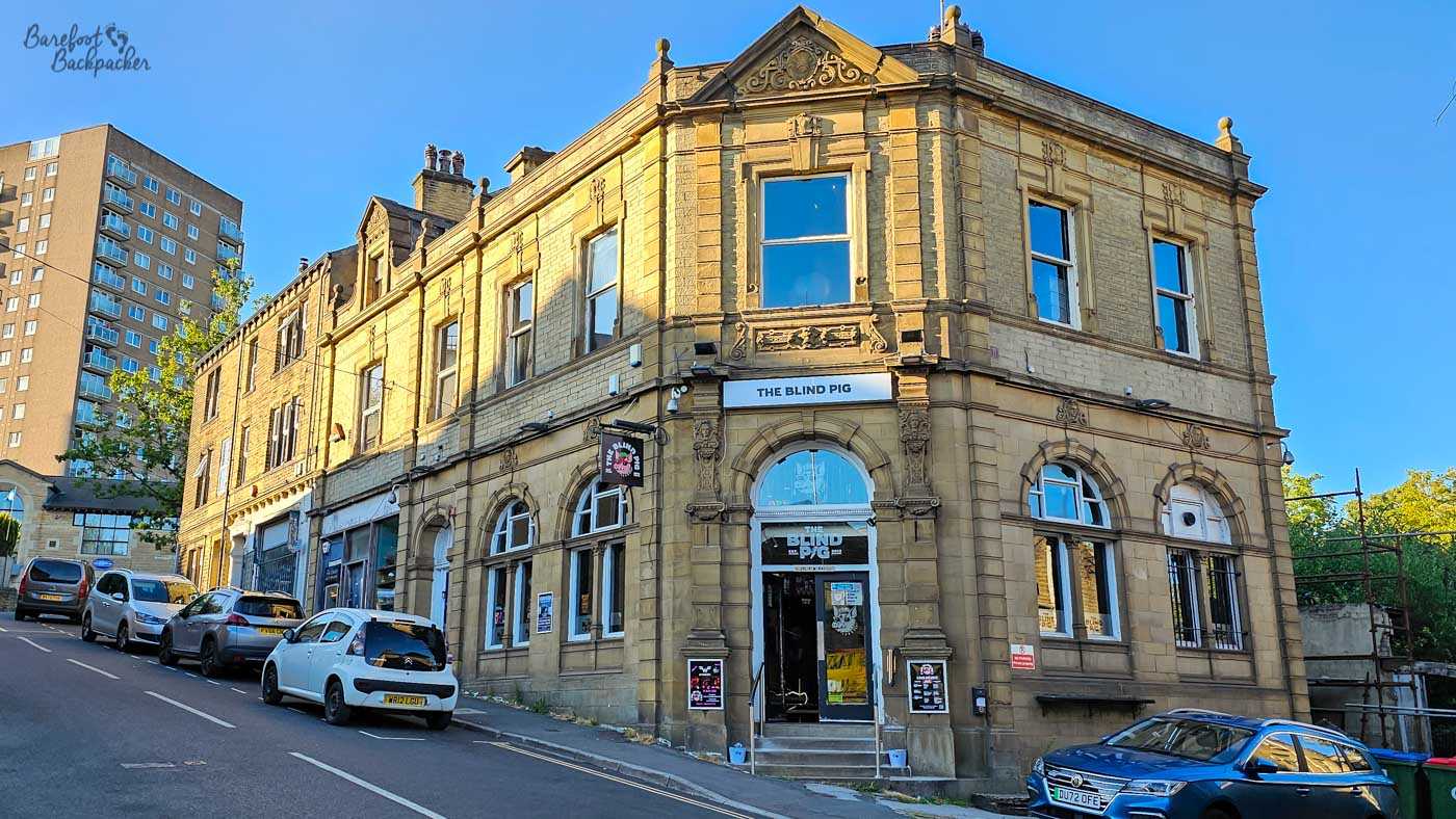 The Blind Pig occupies an imposing Victorian corner building of honey-coloured stone, its ornate façade standing on a steep hill with parked cars lining the roadside. Large arched windows and decorative stonework frame the entrance beneath the pub's sign, while modern apartment blocks rise behind the historic building. Bathed in warm evening sunlight beneath a clear blue sky, the pub makes a striking landmark in the centre of Sowerby Bridge.