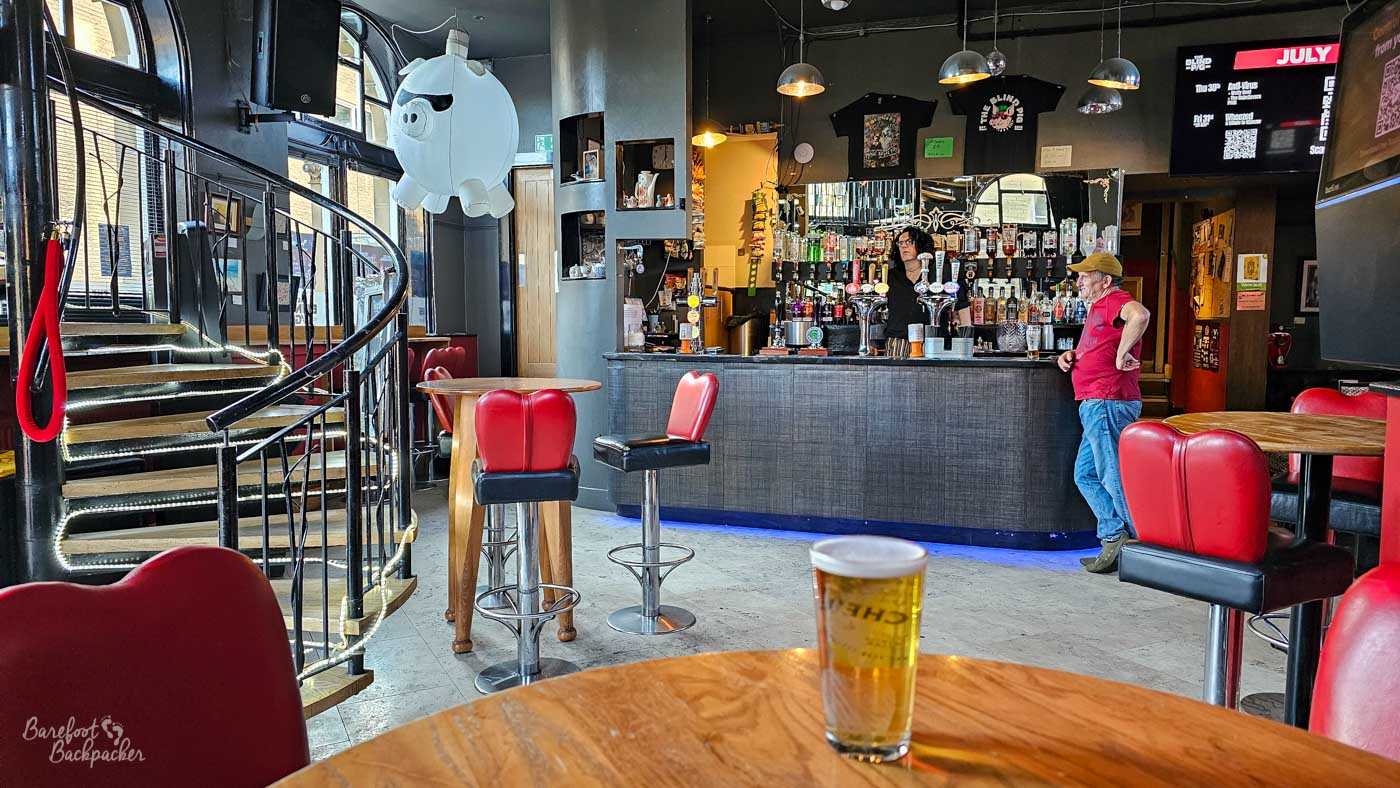 The ground-floor bar combines modern styling with the character of the old building, featuring black walls, polished wooden tables and distinctive red leather bar stools. A sweeping curved staircase rises beside the entrance beneath a giant pig-shaped balloon suspended from the ceiling, while the well-stocked bar sits opposite beneath hanging lights. A pint rests on the table in the foreground as customers chat with the bartender, creating a relaxed early evening atmosphere.
