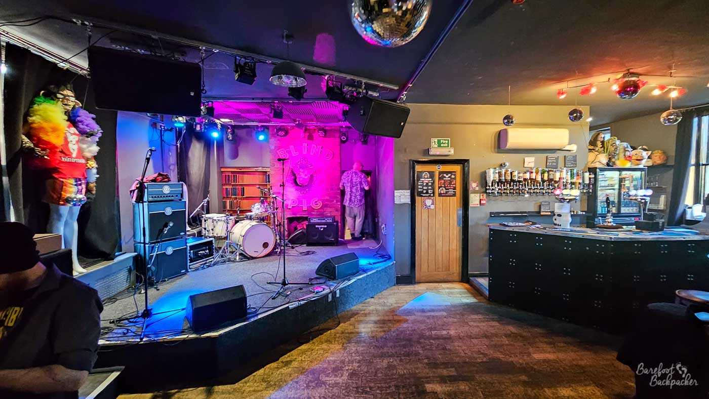 The upstairs room is arranged as an intimate live music venue, with a small stage set beneath colourful purple and blue lighting. Musical equipment including a drum kit, amplifiers and microphones has already been set up while someone makes final preparations before the performance. Disco balls hang from the ceiling and quirky decorations line the walls, giving the venue an energetic atmosphere long before the audience arrives.