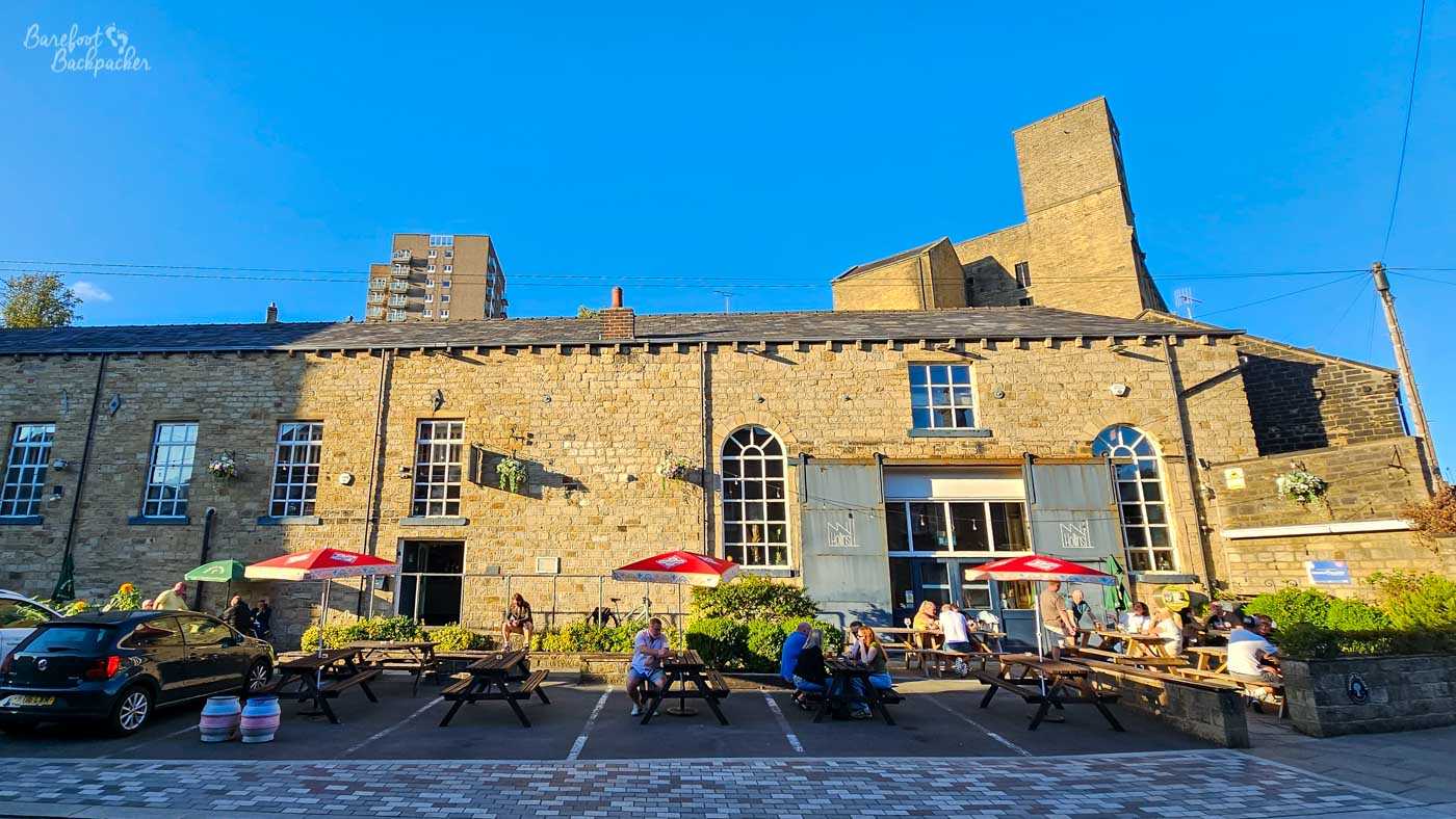 A large former mill building has been converted into a pub, its long stone frontage overlooking a car park filled with wooden picnic benches and red-and-white parasols where groups of people sit enjoying the late afternoon sunshine. Tall arched windows and industrial-style doors hint at the building's past, while modern apartment blocks rise behind the roofline. The warm golden light catches the honey-coloured stone, contrasting with a vivid blue sky and giving the whole scene a relaxed, summery atmosphere.