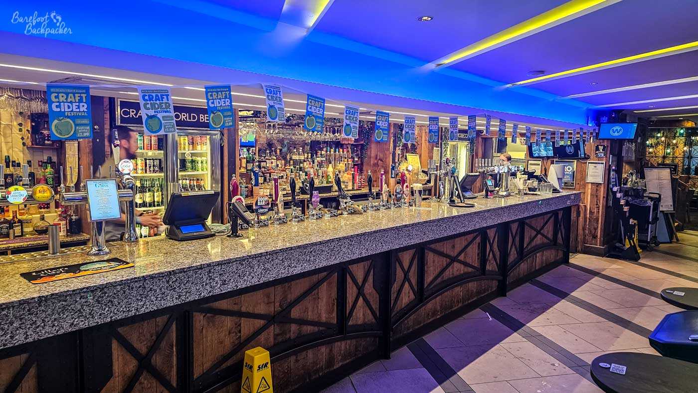 A long granite-topped bar dominates the room beneath blue accent lighting and rows of colourful craft cider festival banners hanging from the ceiling. Fridges stocked with bottled beers line the back wall alongside hand pumps and spirits, while televisions and seating areas extend into the distance. The combination of modern lighting and traditional timber panelling gives the pub a contemporary yet familiar feel.