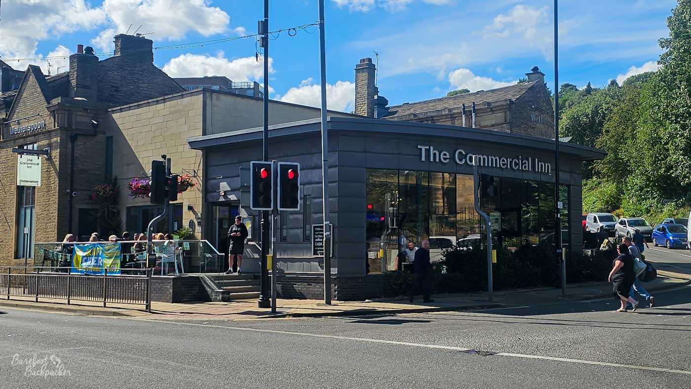 The Commercial Inn stands prominently on a busy road junction, combining an older stone building with a striking modern glass-fronted extension displaying the pub's name. Outdoor seating occupies a raised terrace beside the entrance where customers enjoy the sunshine, while traffic lights, pedestrians and passing cars emphasise its central position within Sowerby Bridge.