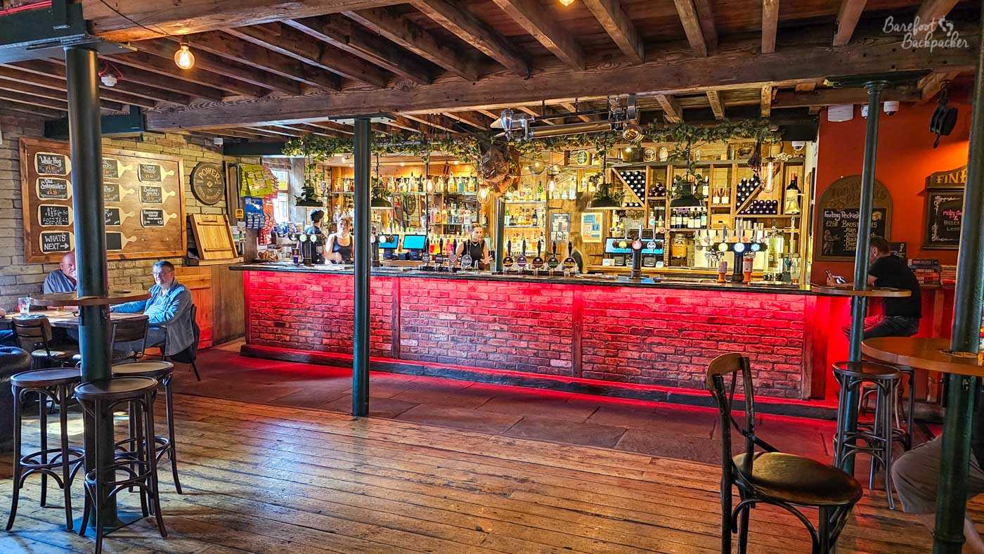 Inside, exposed timber beams stretch across the ceiling above an expansive open room centred on a striking brick-fronted bar illuminated with deep red lighting beneath the counter. Shelves of spirits, cask ales and wines fill the back bar while customers relax at tables around the spacious interior. The blend of rustic timber, exposed brick and colourful lighting creates a lively venue that feels equally suited to casual daytime drinks or busy evening crowds.