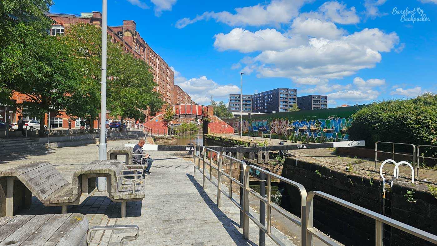 A broad view along an empty canal lock in an urban area, with wooden lock gates, metal railings and benches beside the paved towpath. A man sits on one of the benches, while large red-brick former industrial buildings rise behind trees on the left. Graffiti covers a long wall beyond the lock on the right, with modern apartment blocks in the distance beneath a bright blue, cloud-filled sky.