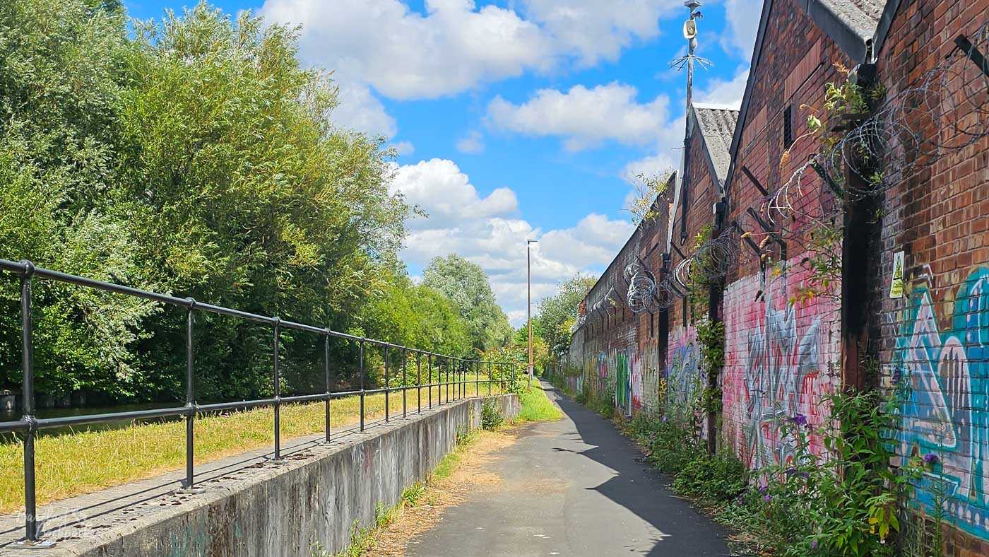 A narrow paved canal towpath running beside a long red-brick industrial building covered in colourful graffiti. Coils of razor wire line the upper walls of the building on the right, while dense green trees and vegetation screen the canal on the left behind black metal railings. The straight path disappears into the distance beneath a bright blue summer sky.