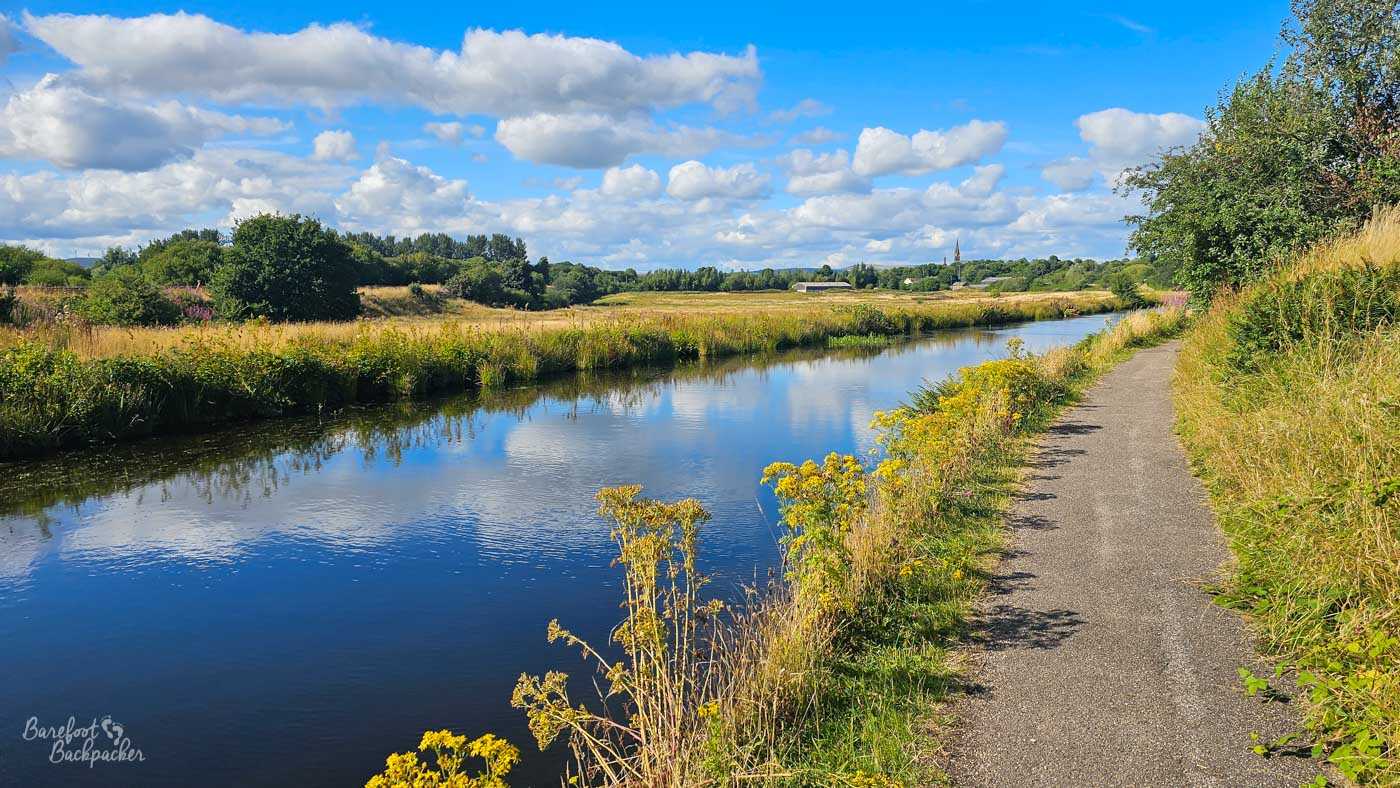 A wide, open stretch of canal through grassy countryside, with a paved towpath following the right bank. Tall grasses and yellow wildflowers grow beside the water, with fields, scattered trees and a few distant buildings beyond. The blue water reflects a bright summer sky filled with large white clouds.