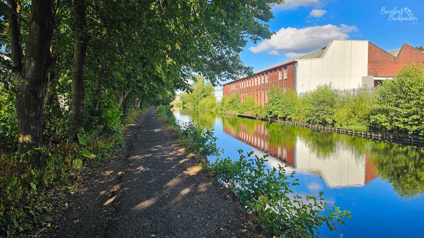 A shaded towpath running alongside the canal, with mature trees forming a leafy canopy on the left. Across the water stands a long red-brick industrial building with a large pale-clad section, reflected clearly in the canal below. Bushes and other vegetation grow thickly along the opposite bank under a bright blue sky.