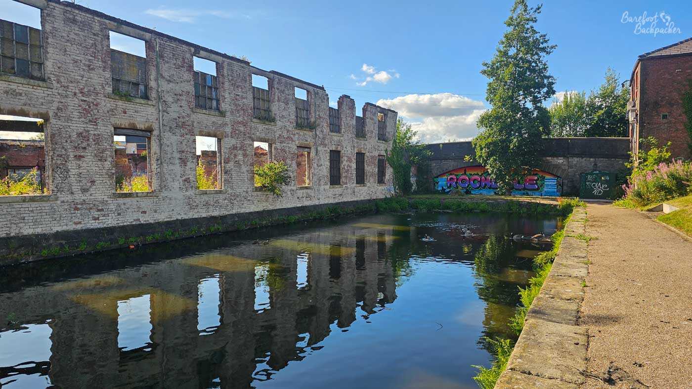 A canal passing a roofless, derelict industrial building with long white-painted brick walls and rows of empty window openings. The ruined building is reflected in the dark water, while the towpath runs along the opposite bank on the right. Ahead, a stone bridge carries the colourful ROCHDALE mural seen in a previous image, partly obscured by a tree.