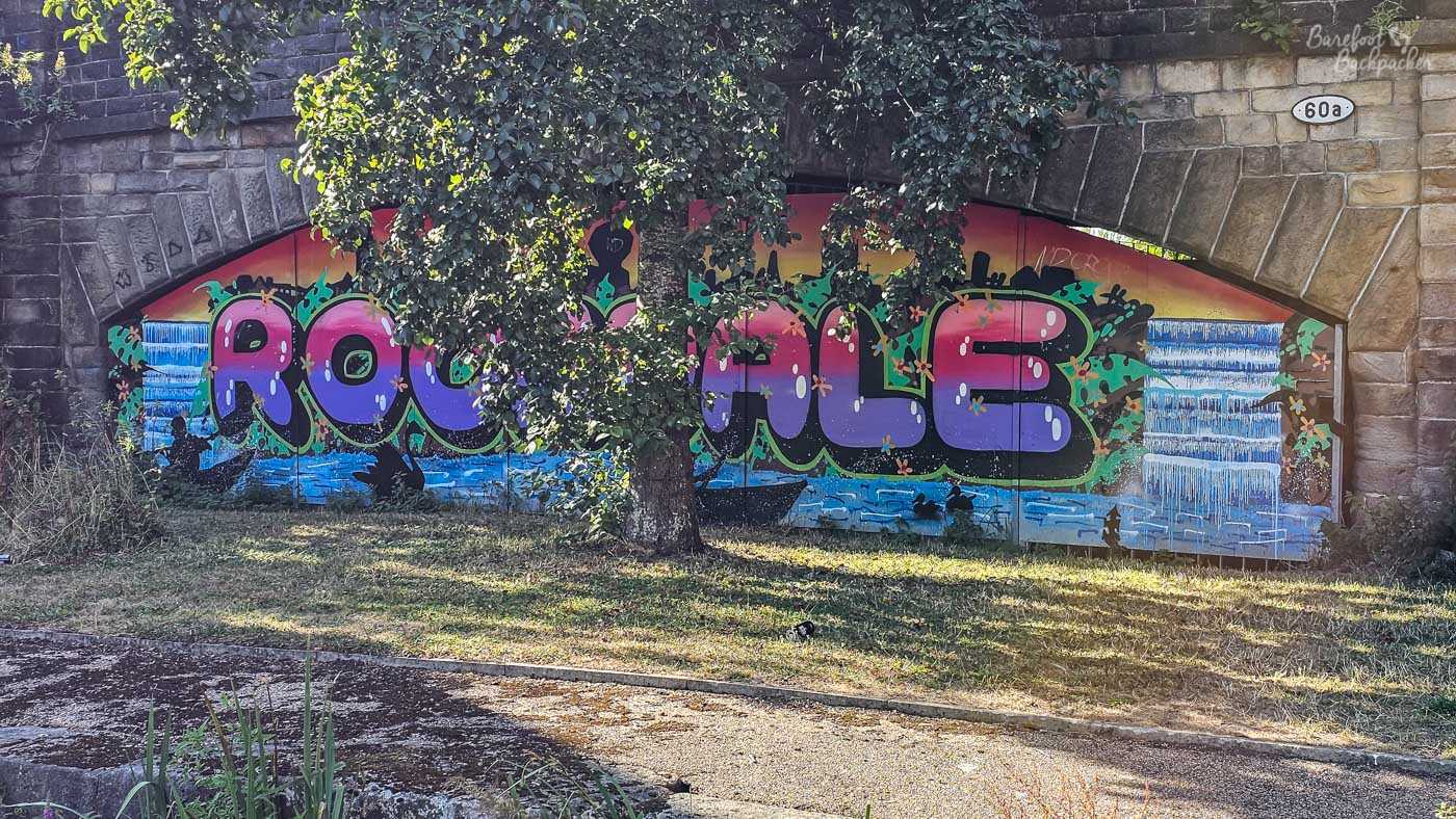 A large colourful graffiti mural beneath a broad stone railway arch beside the canal. Huge rounded purple and pink letters spelling ROCHDALE stretch across the wall, surrounded by green foliage designs and painted blue water and waterfalls. A mature tree partly obscures the centre of the mural, with the stone arch and a small 60a marker visible above it.