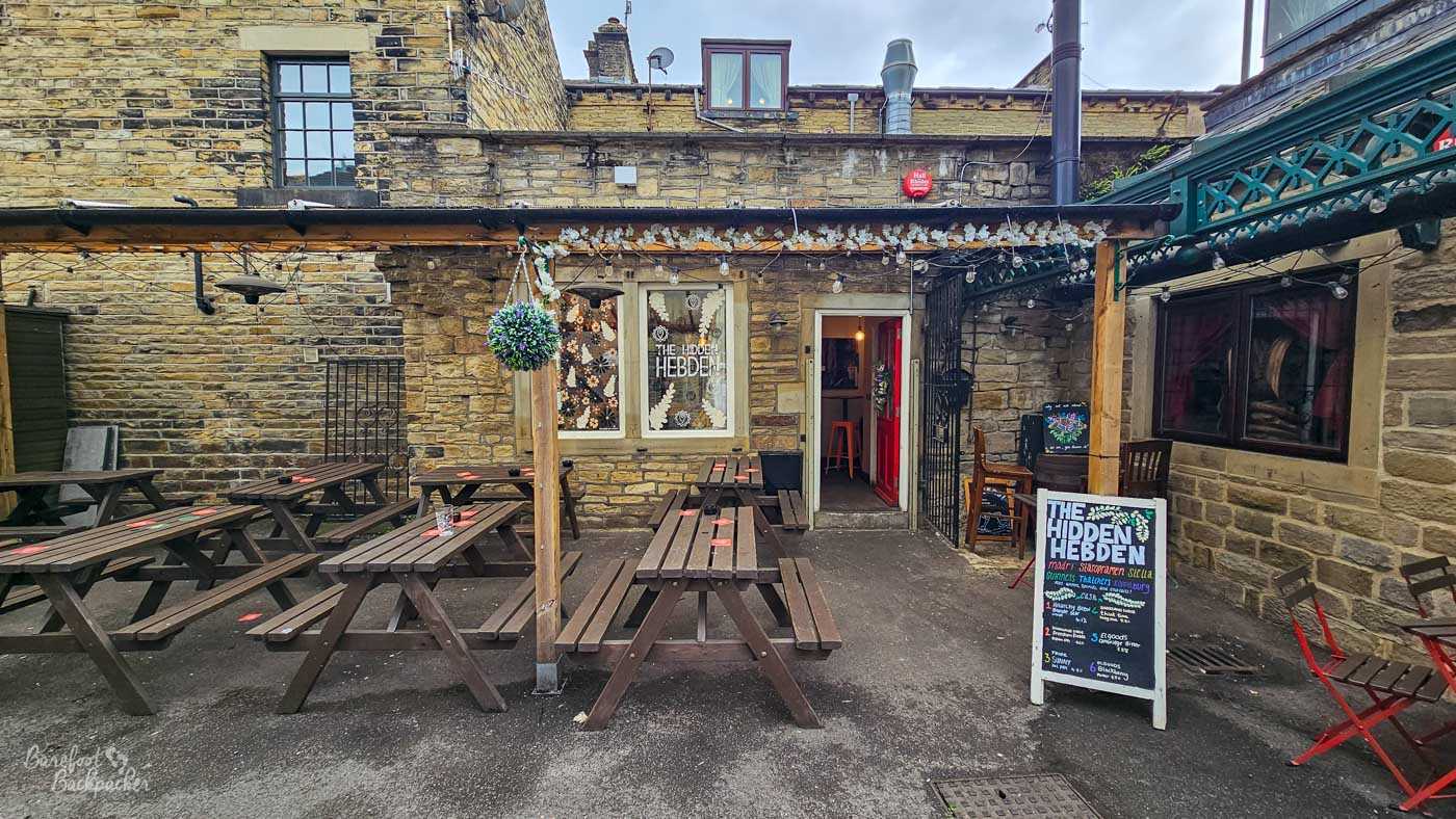 The secluded outdoor courtyard of The Hidden Hebden in Hebden Bridge, enclosed by old honey-coloured stone buildings. Rows of dark wooden picnic tables fill the yard beneath strings of lights and floral decorations, with the pub entrance set into the stone wall at the rear. “The Hidden Hebden” is written on the window beside the open doorway and again on a freestanding chalkboard advertising drinks. A decorative green-painted iron structure runs along the upper right of the courtyard.