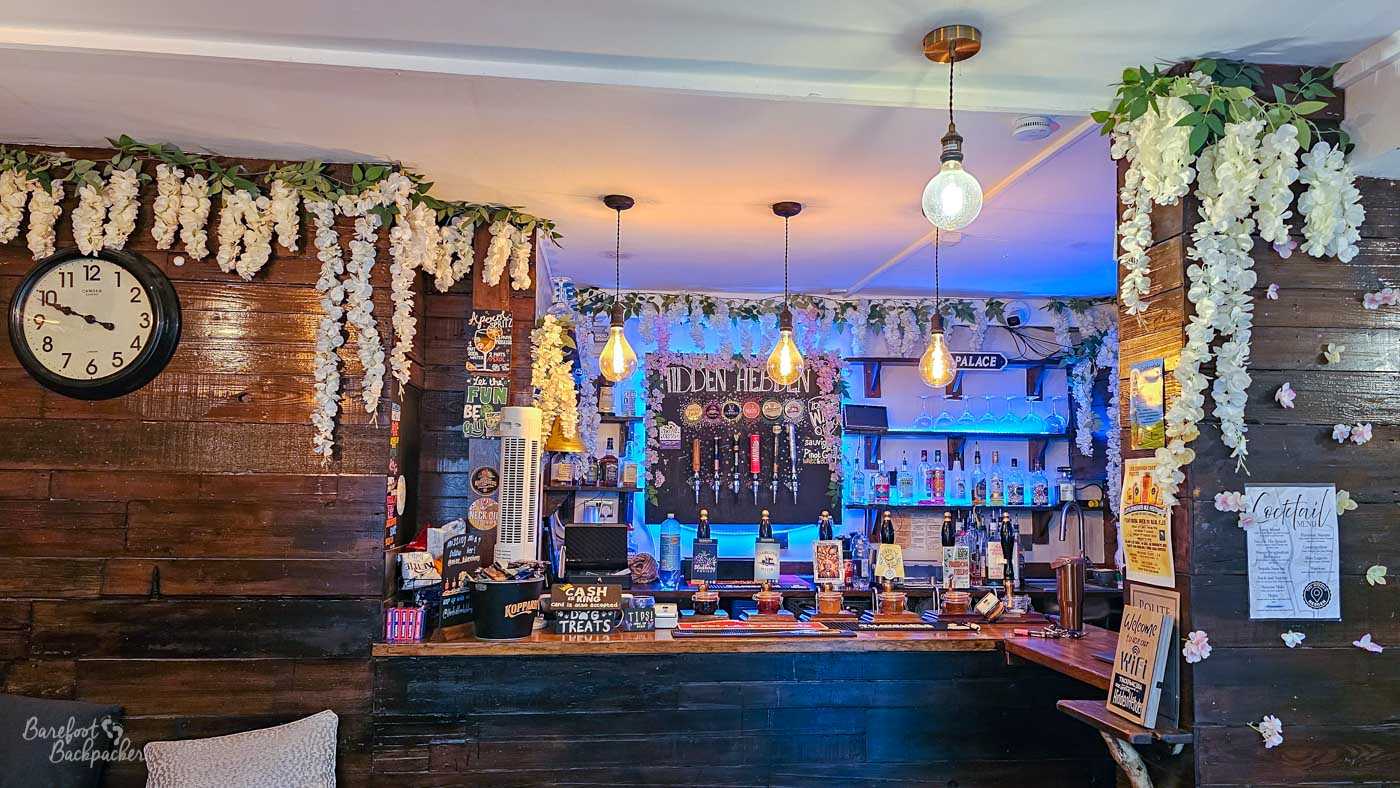 Front view of the compact bar inside The Hidden Hebden, surrounded by dark wooden panelling and cascades of artificial white flowers and greenery. Several hand-pulls and beer taps line the wooden counter, with bottles of spirits displayed on illuminated shelves behind and a chalkboard bearing the pub’s name above them. Warm pendant bulbs hang from the ceiling while blue lighting behind the bar adds a colourful contrast to the otherwise rustic interior.